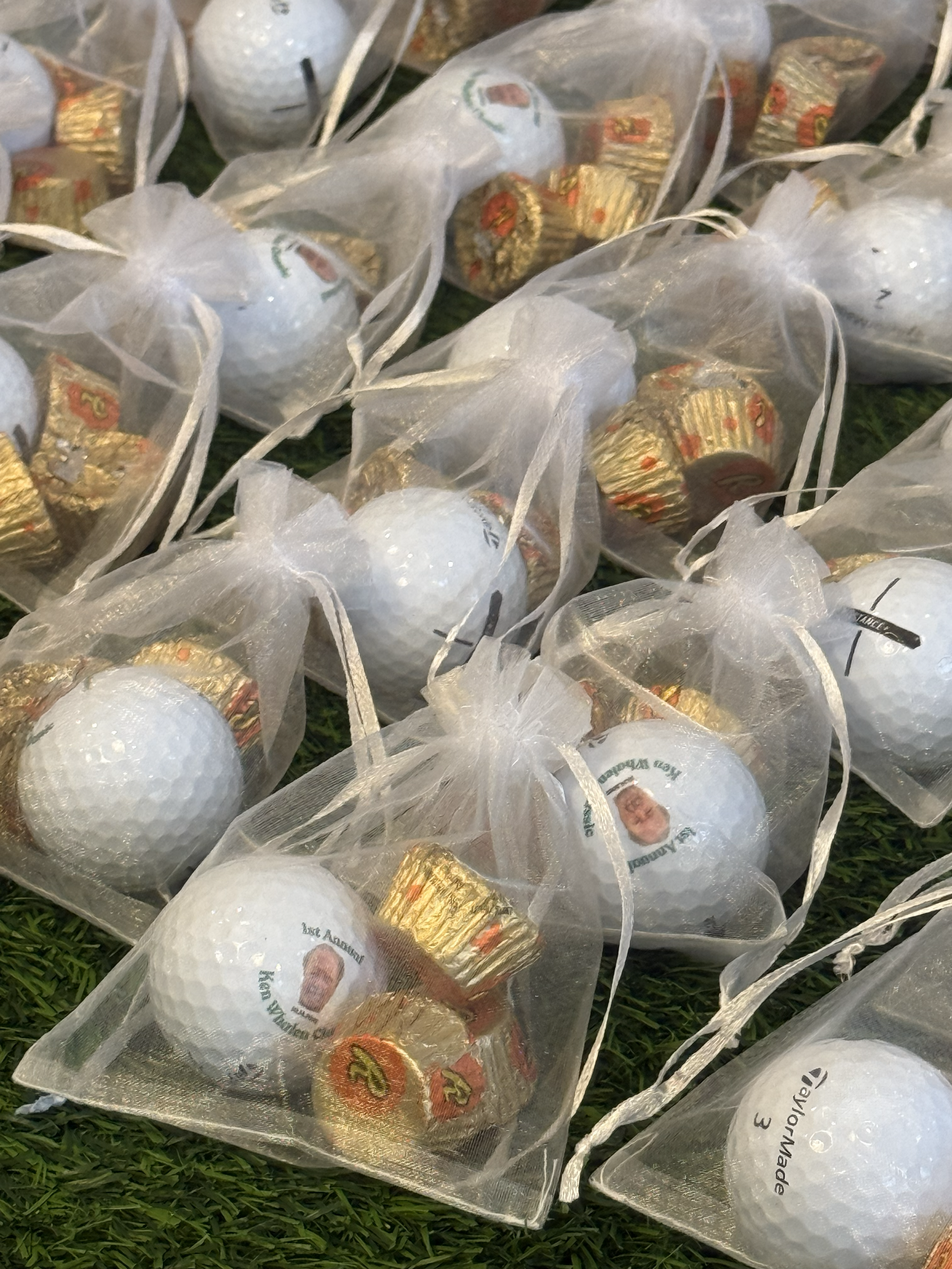 Several white golf balls in clear plastic bags with gold-wrapped chocolates inside, on grass. Some bags feature a photo and name, likely for a memorial or tribute event.