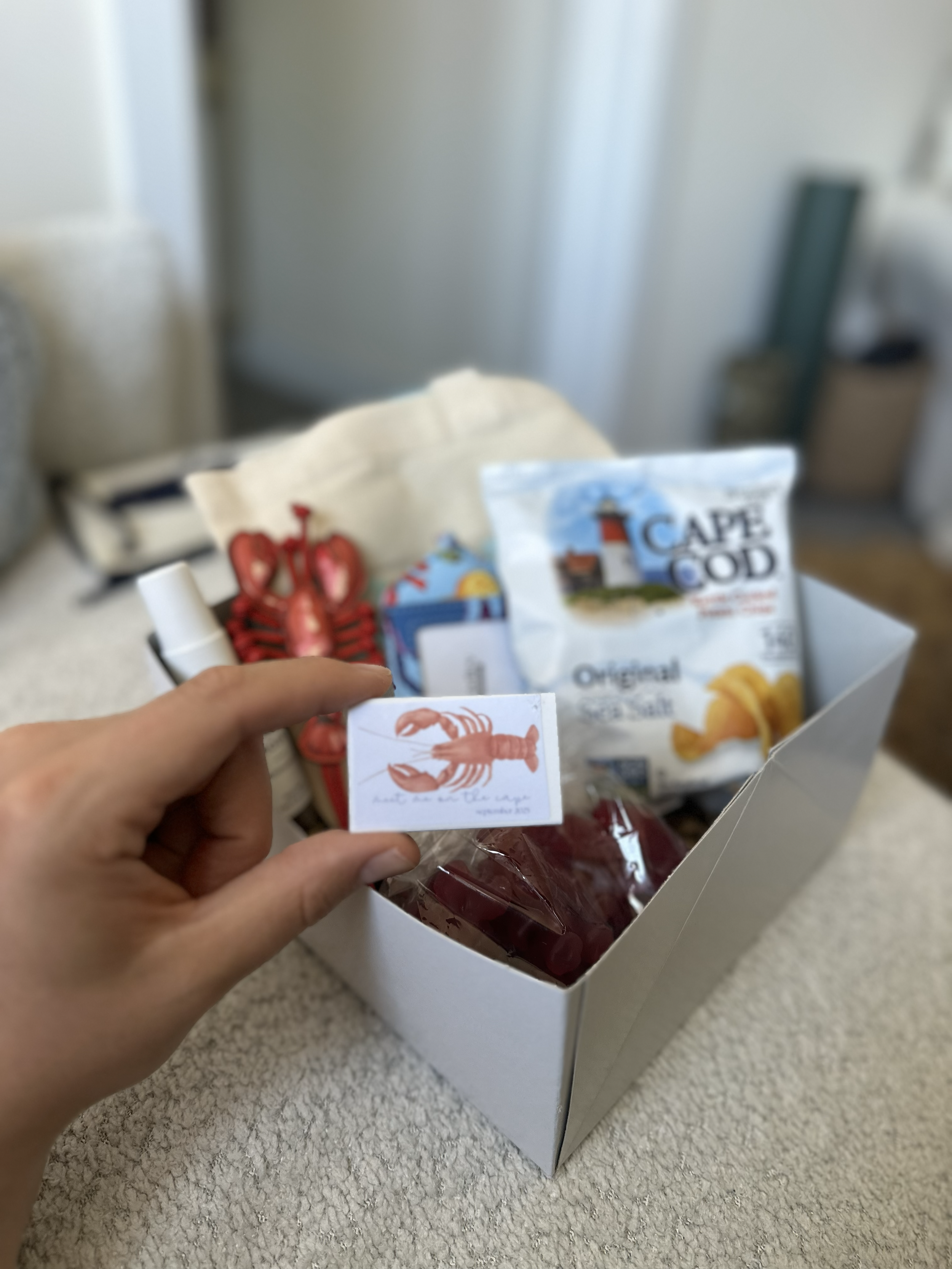 A white box containing various souvenirs and snacks, including a red lobster-shaped item, a bag of Cape Cod Original Chips, and a small cylindrical container, with a person's hand holding a small card featuring a lobster illustration.