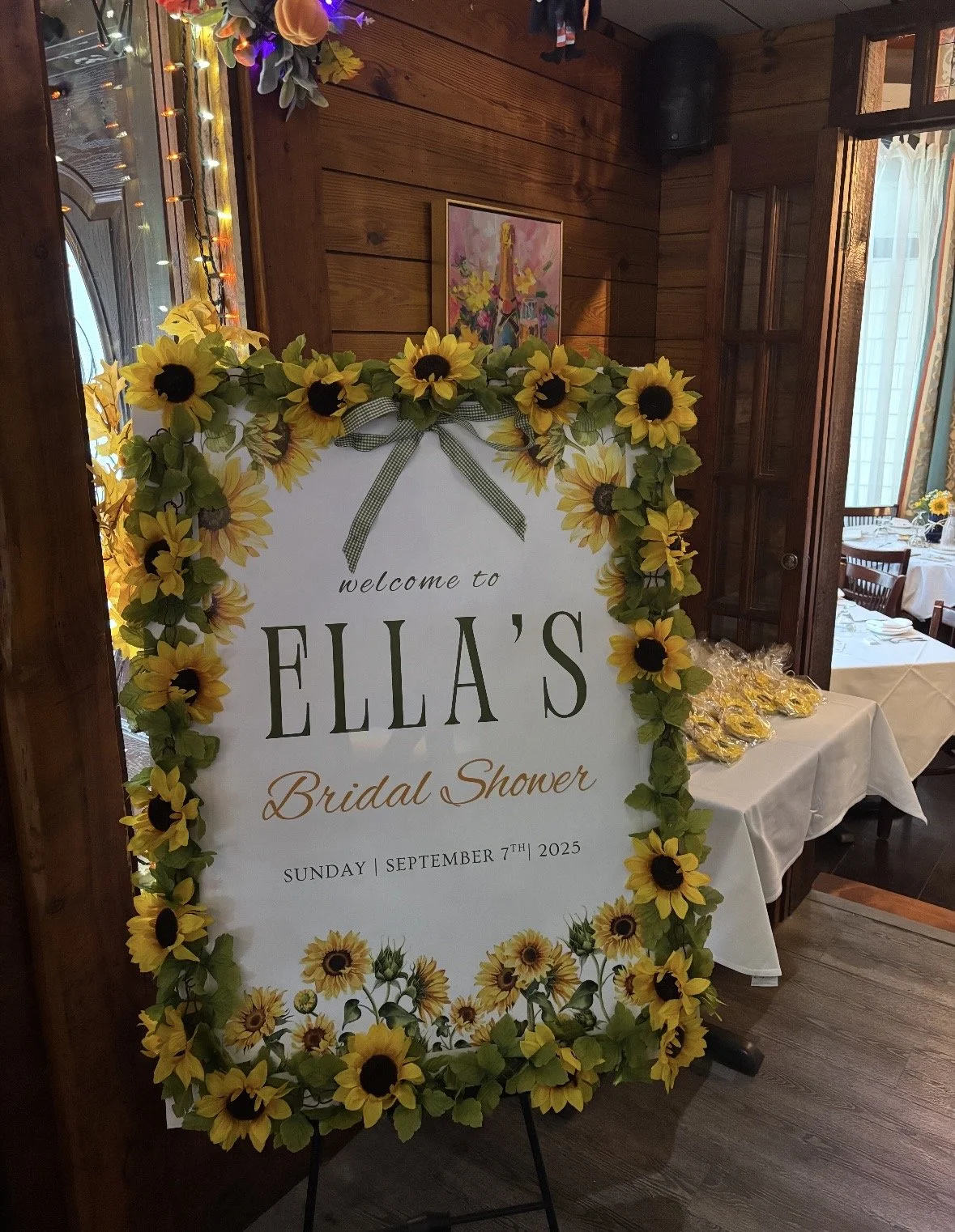 A welcome sign for Ella's bridal shower, decorated with sunflowers and a green gingham ribbon, on an easel in a warmly lit room with wooden walls and tables set for a celebration.