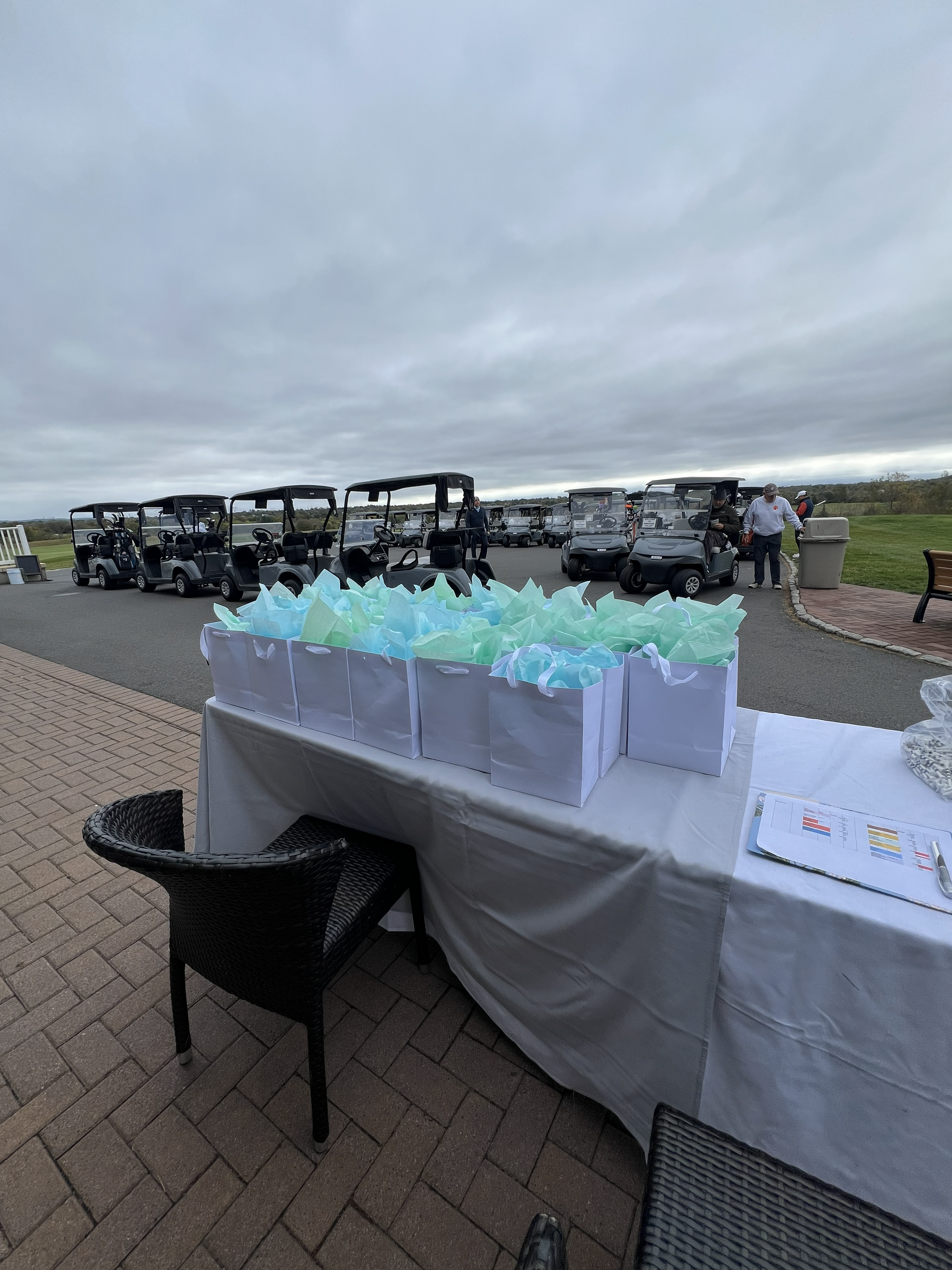 Outdoor scene at a golf course with a table of gift bags in the foreground and golf carts parked in the background under cloudy skies.