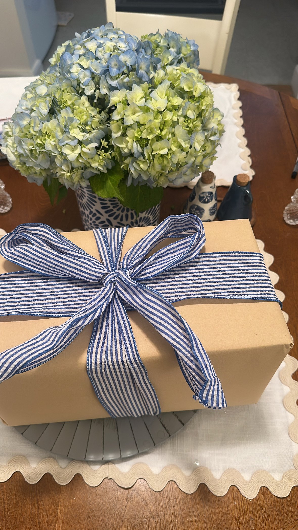 A wrapped gift box with a blue and white striped ribbon on a dining table in front of a vase of light green and blue hydrangea flowers.