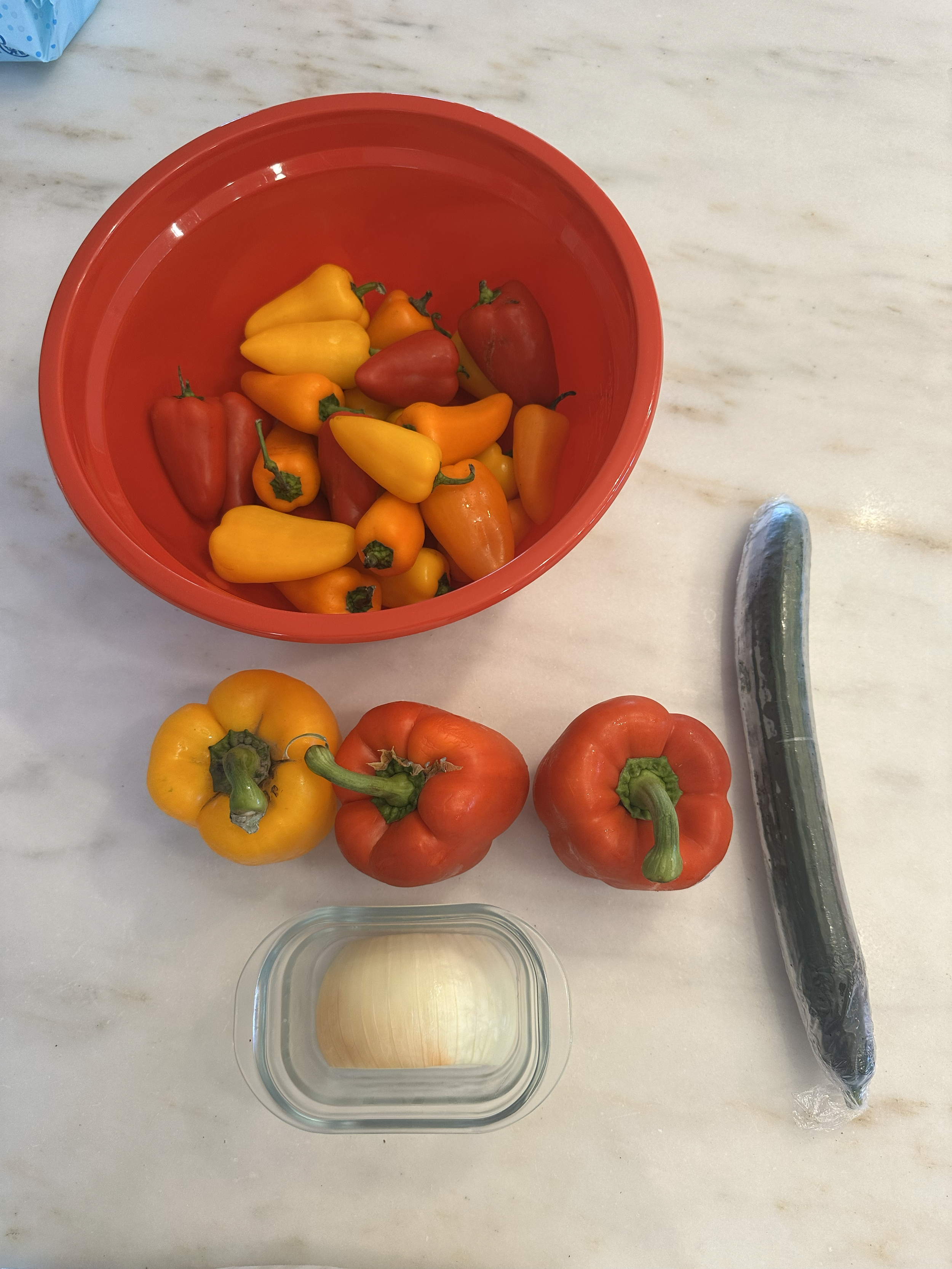 A red bowl filled with multicolored mini bell peppers on a white countertop, with three large bell peppers, a whole onion in a glass container, and a cucumber wrapped in plastic on the surface.
