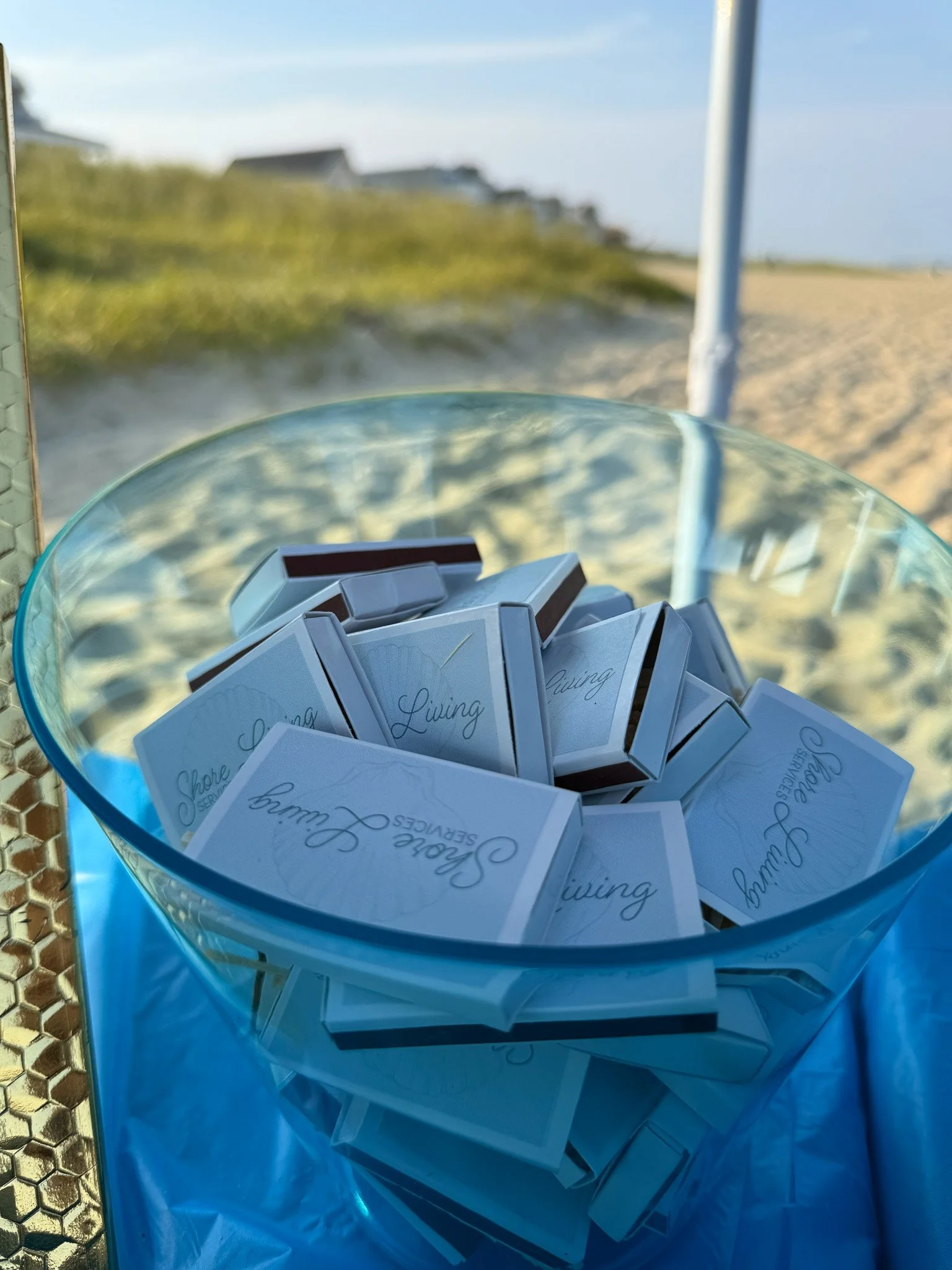 A clear glass bowl filled with folded paper sachets labeled 'Shore Living' and 'Seaweed Serums,' situated on a blue surface with a beach background, including sand, grass, and a blue sky.