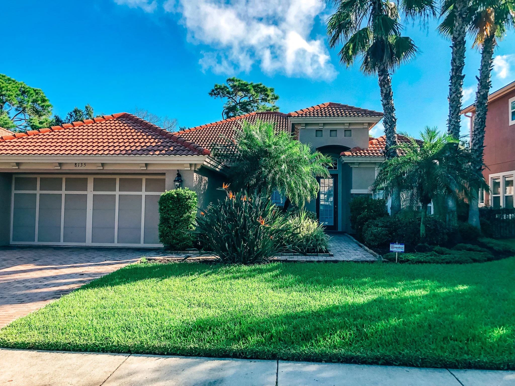 A white house with a brown roof and brick chimney, situated under a cloudy sky, with green foliage on the right side.