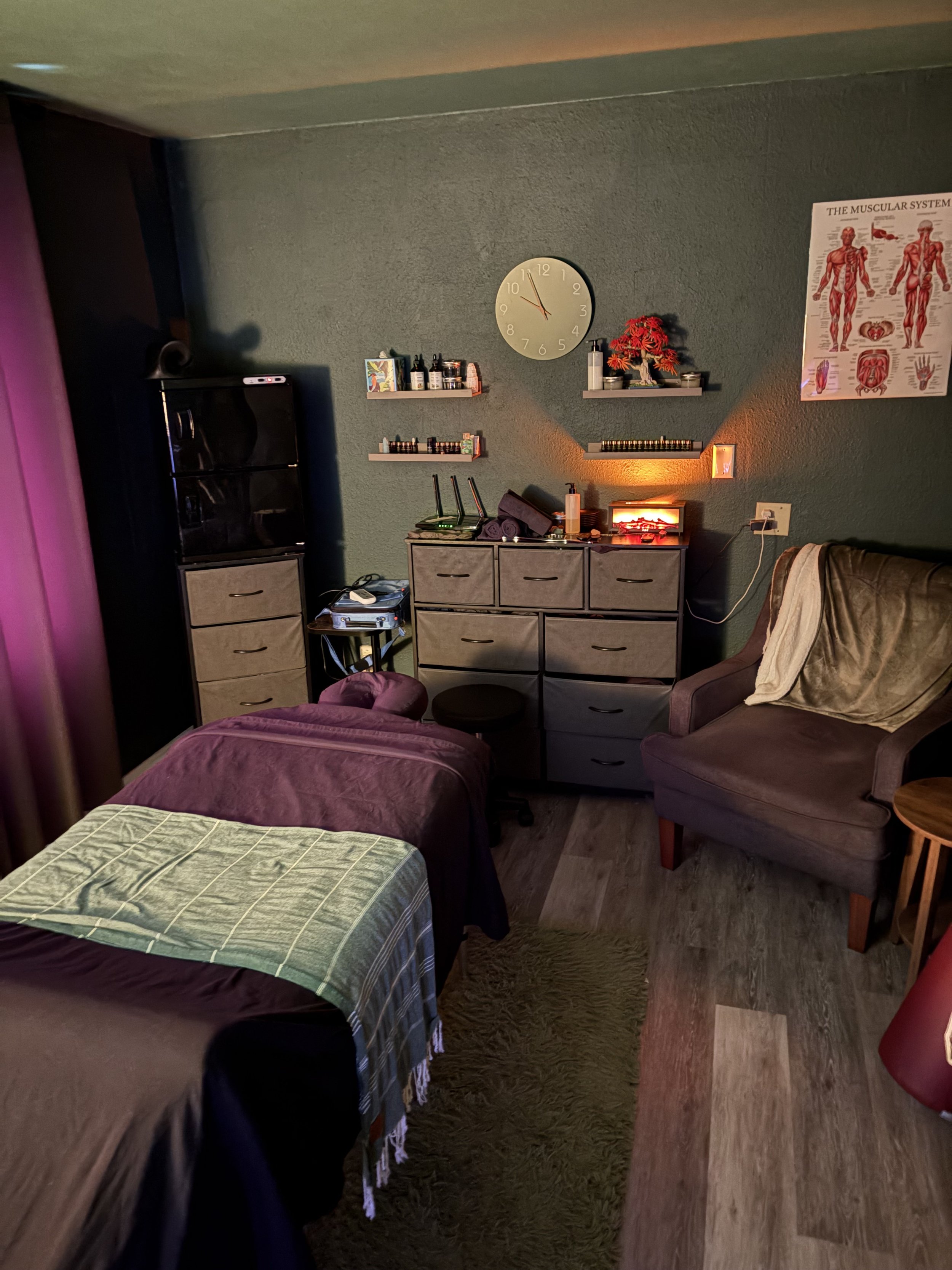 A massage room featuring a massage table with a checked blanket, a dresser, a green shag rug, a gray armchair with a blanket, a small side table, and wall decorations including a clock and a health chart.  Room is designed for men to keep them calm a
