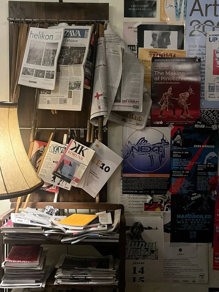 A cluttered room corner with a wooden newspaper rack holding newspapers and magazines, a lamp with a beige shade, a shelf filled with papers and folders, and a wall with various posters and flyers.
