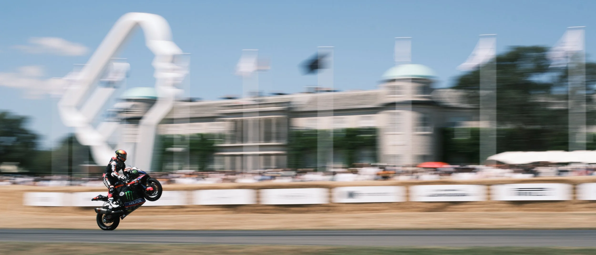 A motorcycle racer performing a wheelie on a race track with a blurred background of spectators, flags, and buildings.