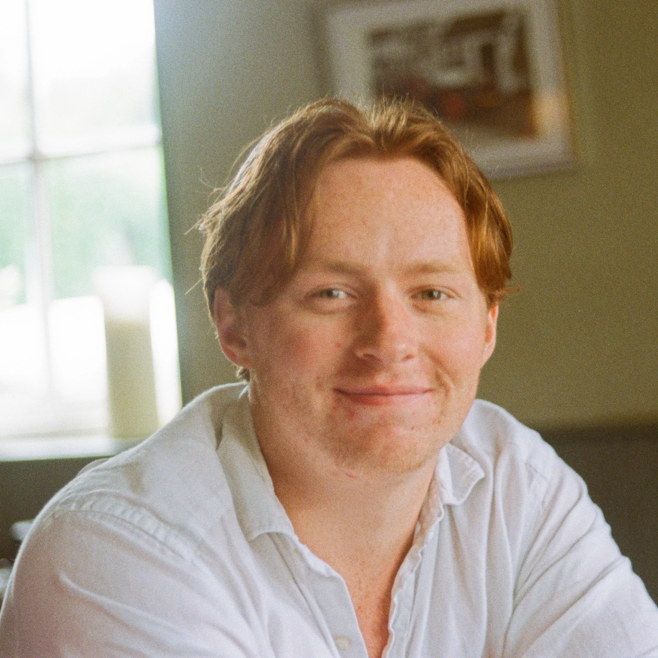 A young man with red hair and a white shirt smiling in a warmly lit room.