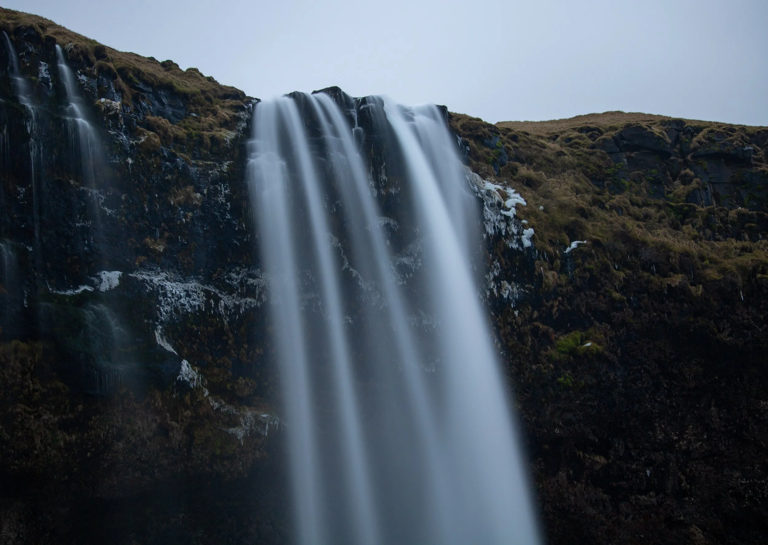 A multi-tiered waterfall cascading down a dark, rocky cliff with patches of snow and moss, overcast sky in the background.