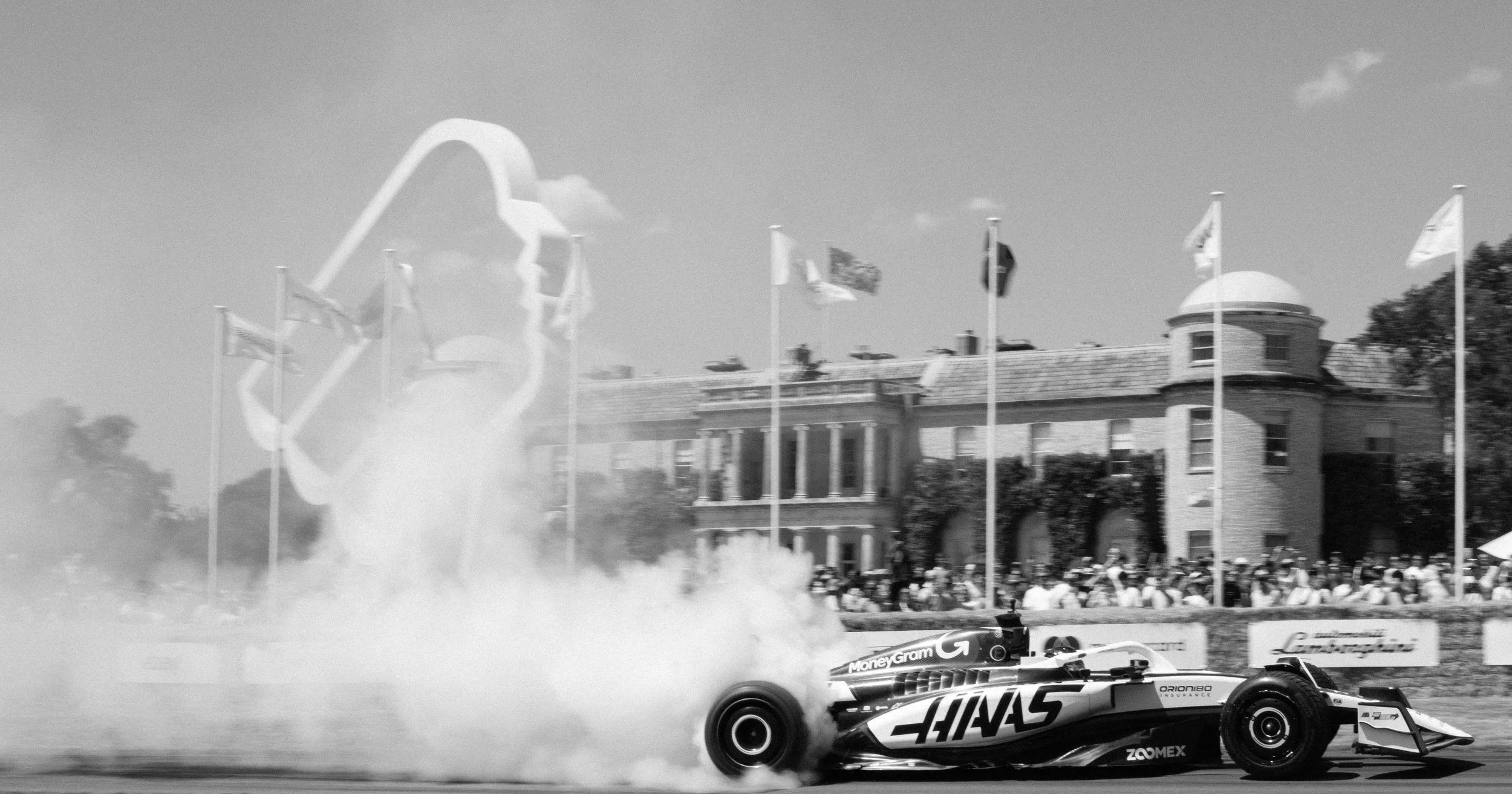 A black and white photo of a Formula 1 car in a burnout at a racing event, with smoke coming from the tires. In the background, there is a grandstand with spectators and a historic building with a dome and flagpoles.