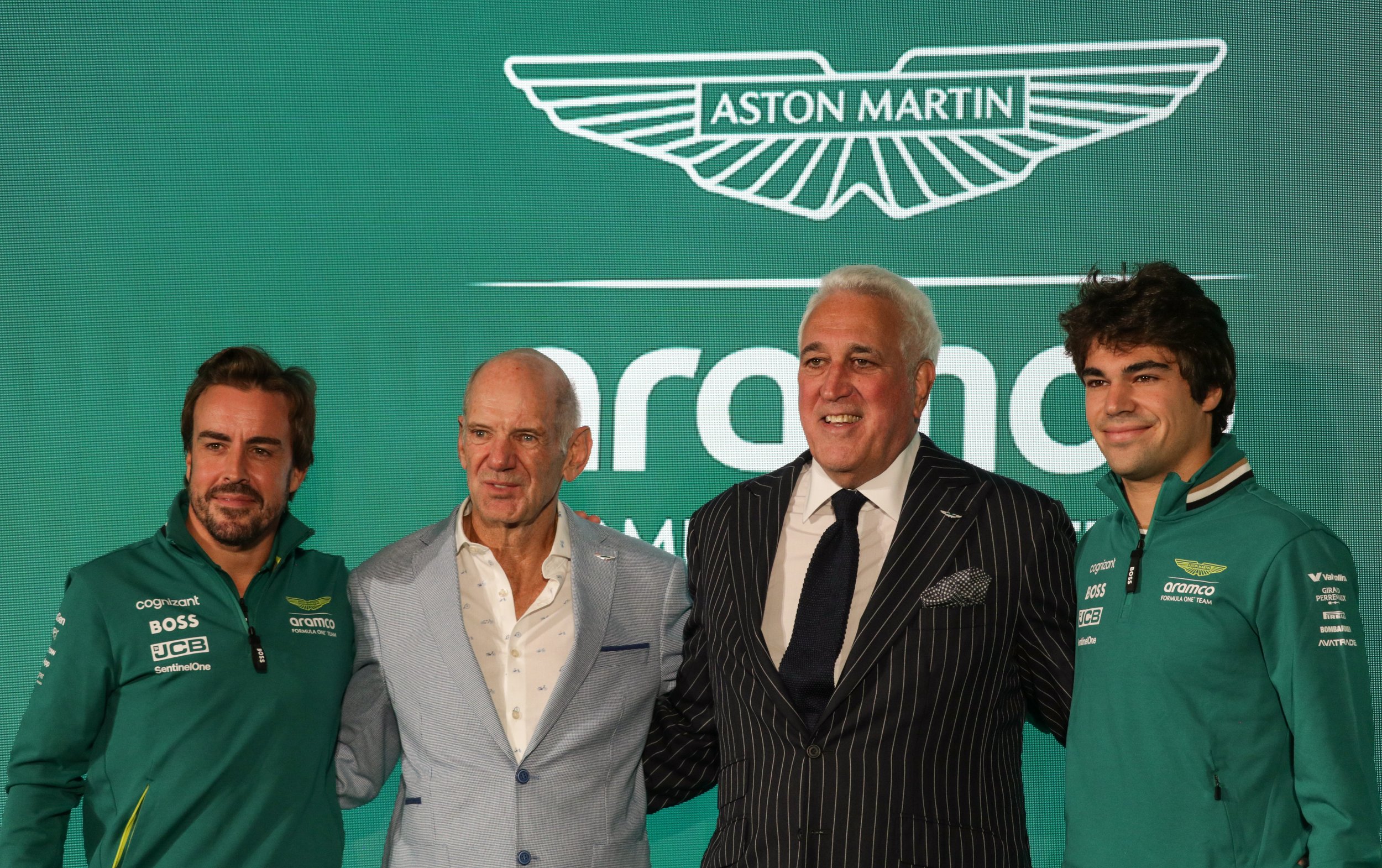 Four men standing together at an event, with a green background displaying the Aston Martin logo and the word 'Aramco.' Two men are wearing green Aston Martin team jackets, while the other two are dressed in suits.