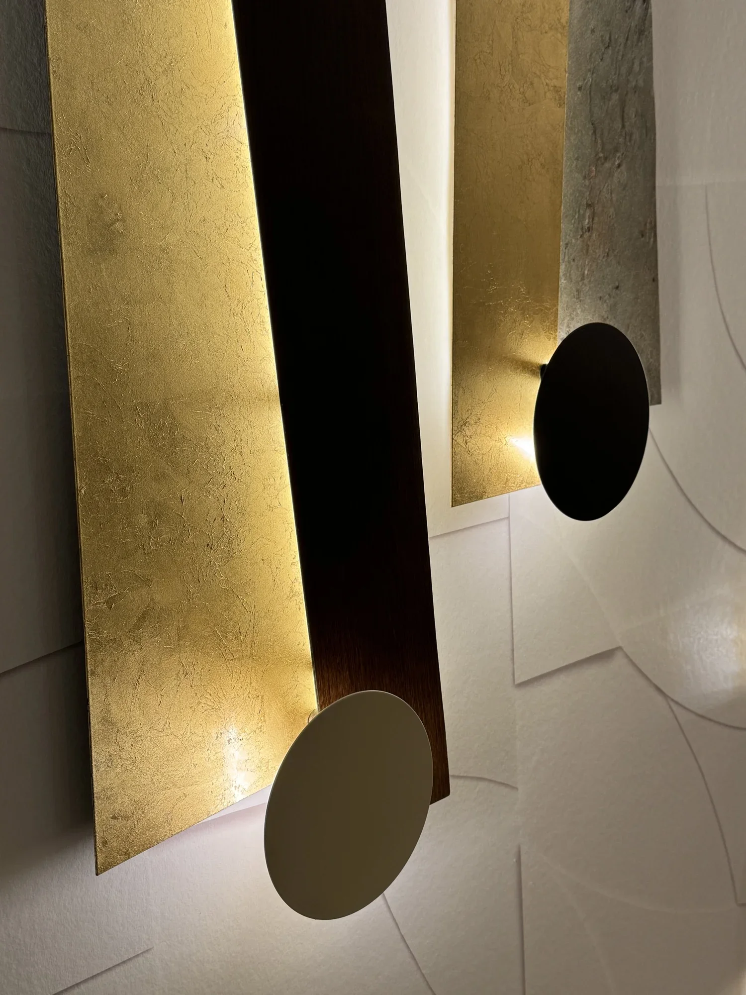 Lampe murale moderne avec plaques rectangulaires en or et en pierre, illumine une pièce avec des lumières intégrées et des cercles noirs et blancs en bâton attachés. La pièce a un mur blanc avec une texture délicate.