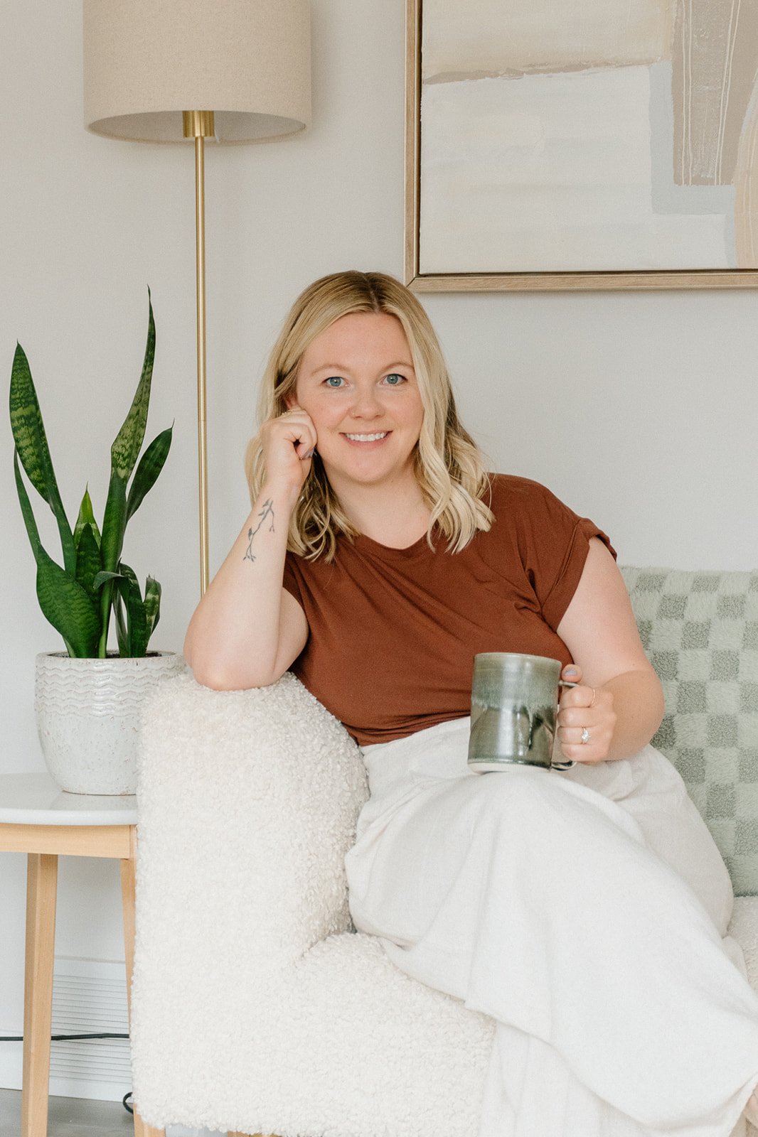 A woman with blonde hair, smiling and sitting comfortably on a cozy, white textured armchair, holding a ceramic mug. She is wearing a brown T-shirt and light-colored pants, with a tattoo on her left arm. A tall potted snake plant is on a side table beside her, and a lamp and abstract artwork are on the wall behind her.