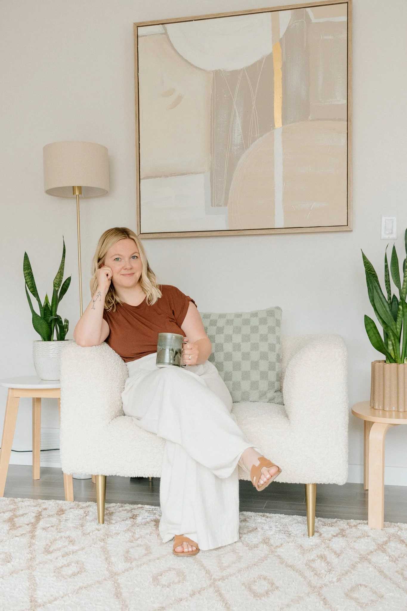 A woman sitting on a cozy white textured sofa, holding a coffee mug, in a modern living room with abstract artwork, potted plants, and neutral-colored decor.