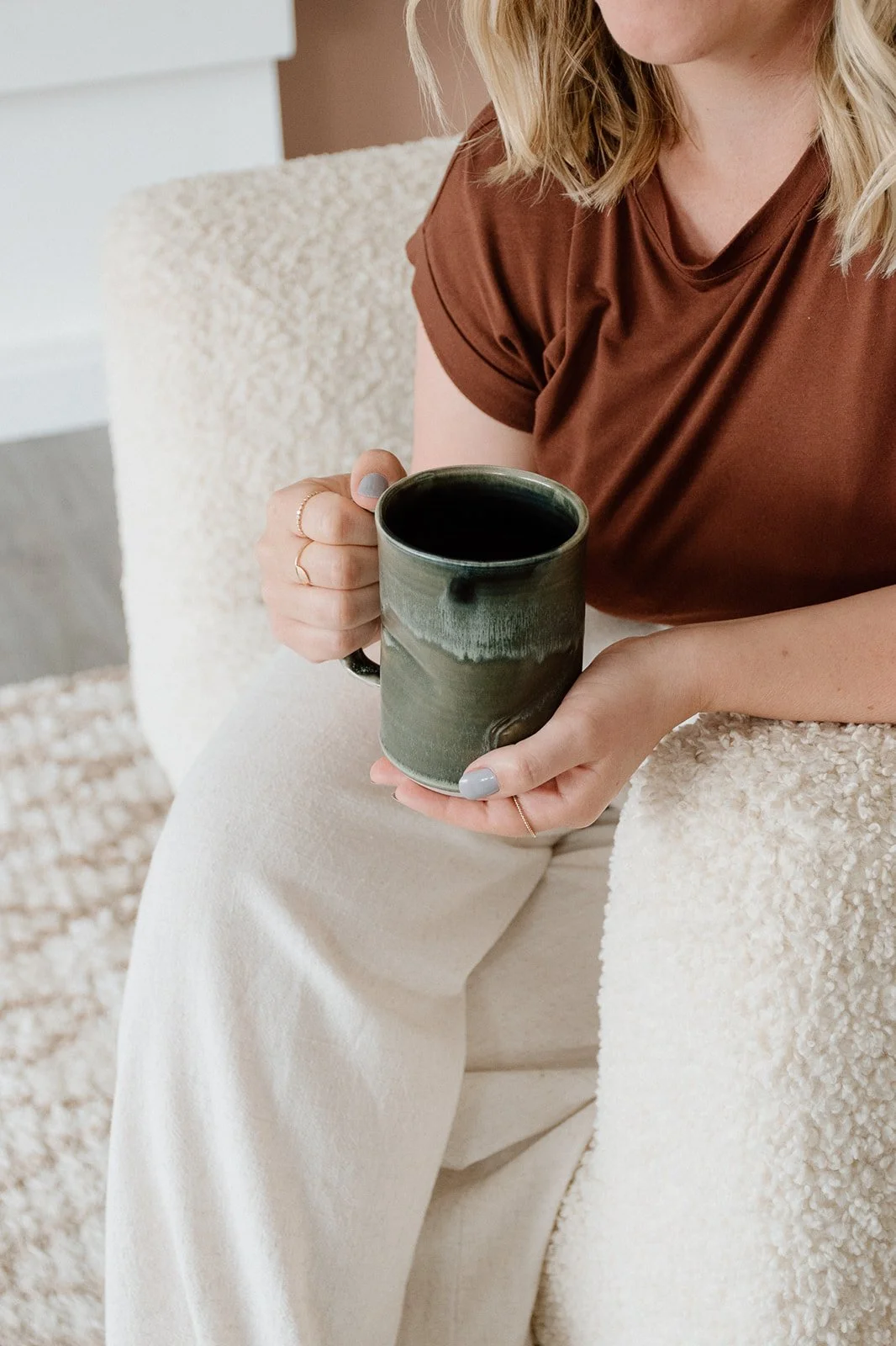 A woman with blonde hair wearing a brown shirt and light-colored pants, holding a dark green ceramic mug while sitting on a cozy, textured cream-colored chair.