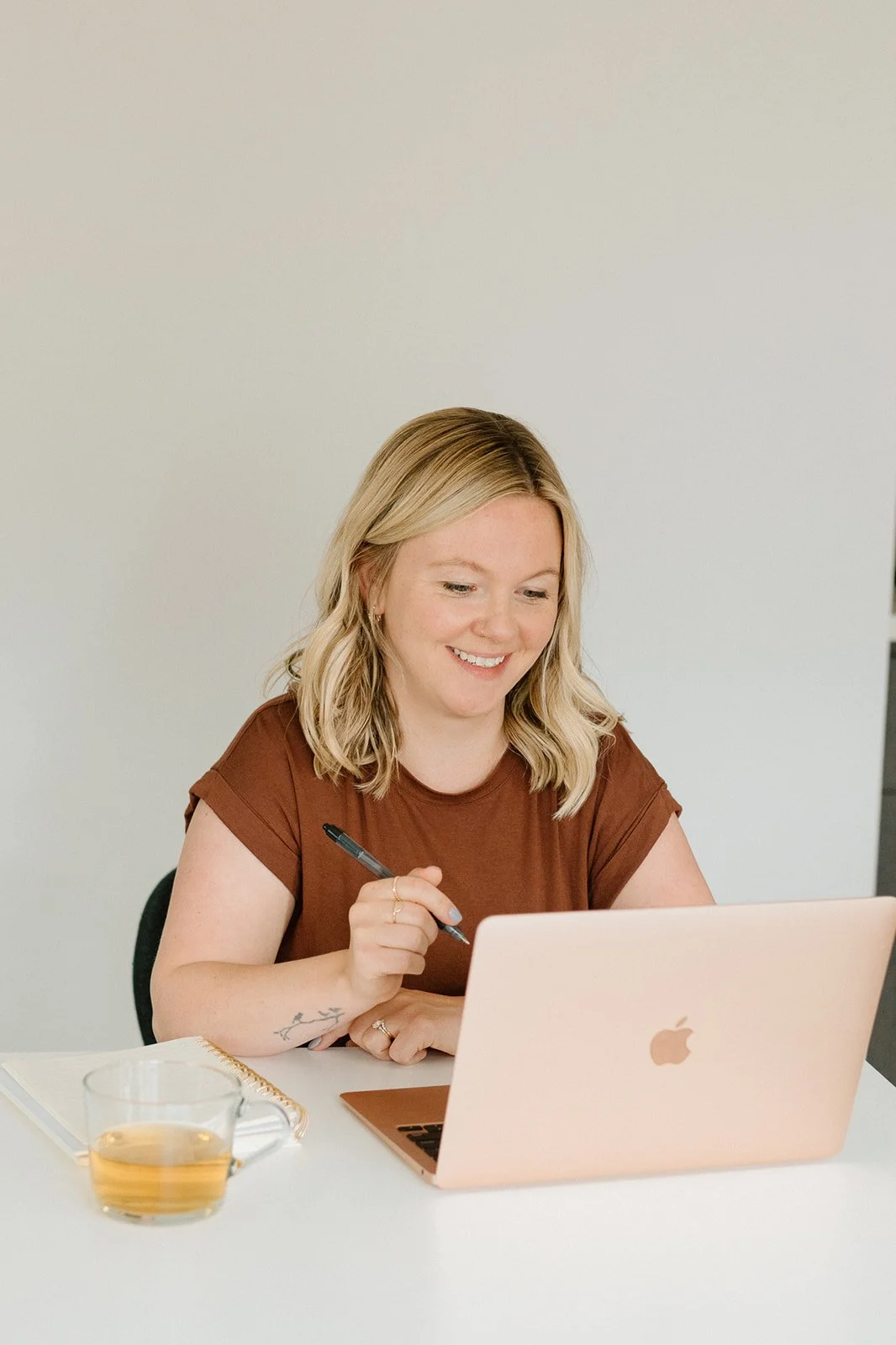 A woman with shoulder-length blond hair and a tattoo on her arm, sitting at a white table, smiling while working on a rose gold MacBook, holding a pen. There is a cup of tea and a notepad on the table.