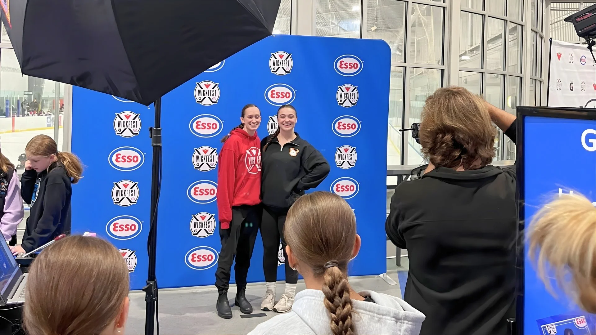 Two women stand in front of a blue background with Wickfest and Esso logos, taking a photo at an indoor event, with several onlookers and photographers capturing the moment.