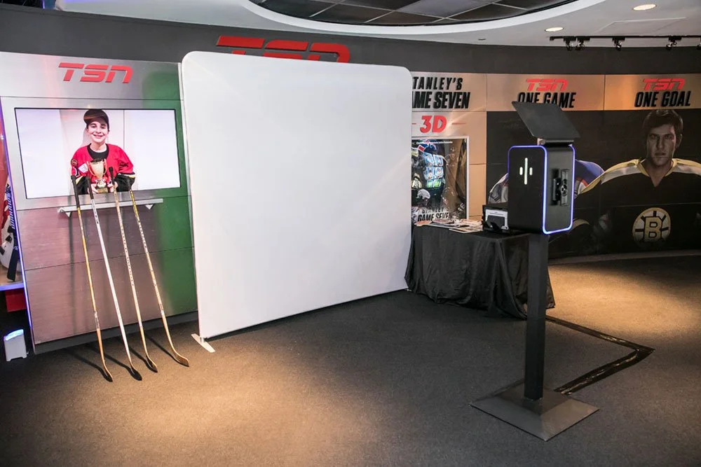An arcade setup with a hockey shooting game featuring a boy on a screen, hockey sticks, a white divider, and a machine on a black stand. The background has hockey-themed posters.
