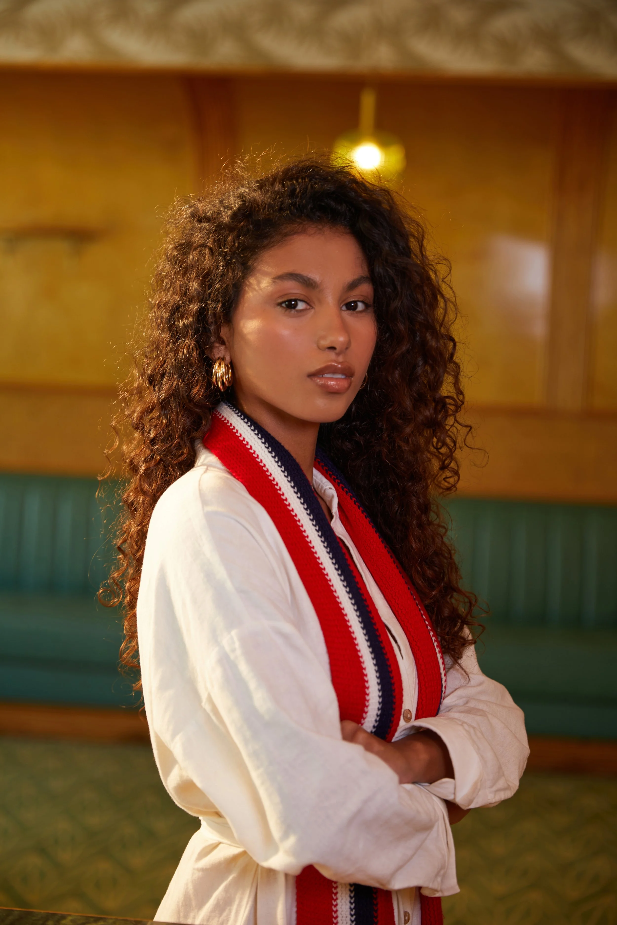 A woman with curly hair wearing a light-colored jacket and a red, white, and blue striped scarf standing with her arms crossed in a warmly lit room.
