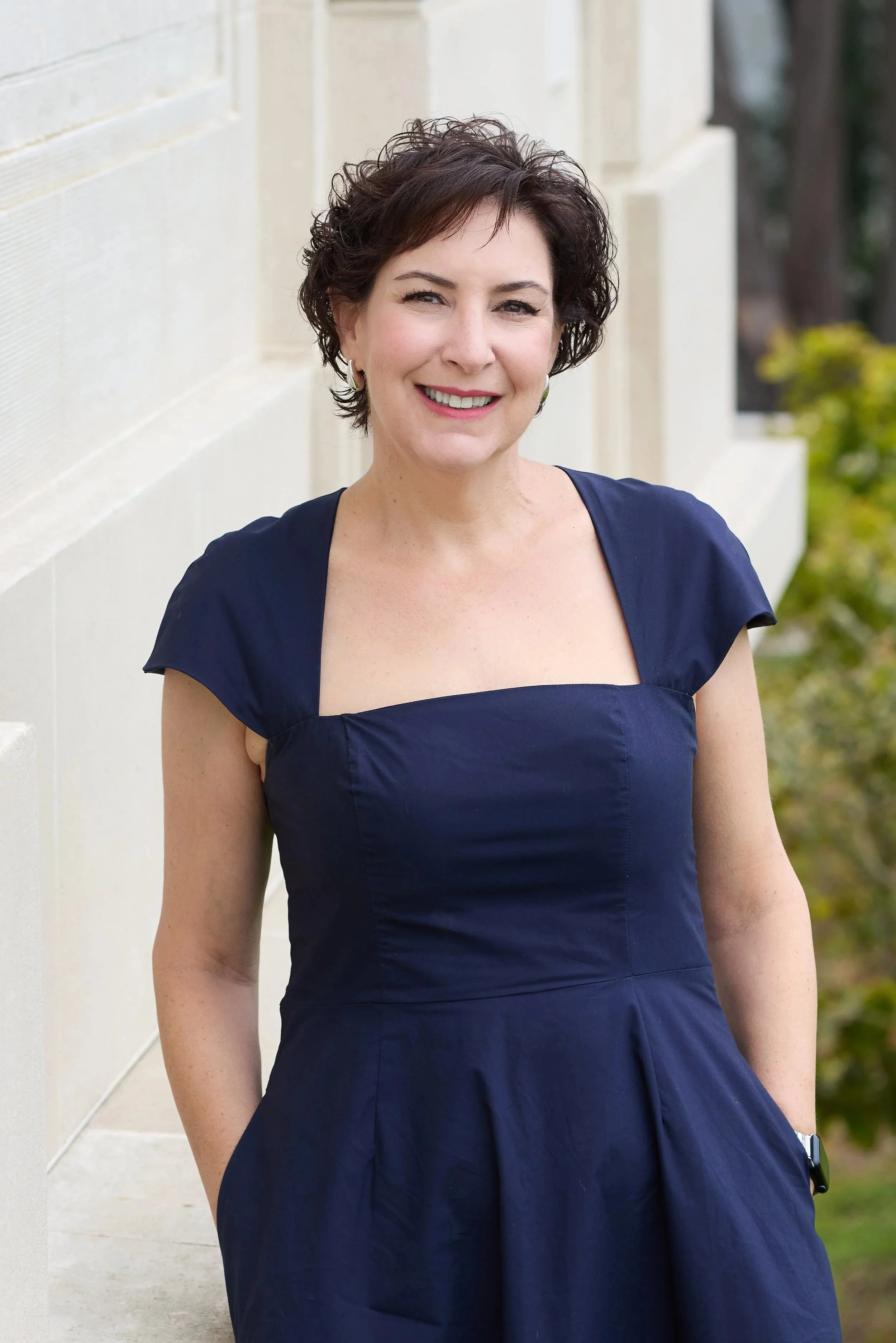 Sarah Spector, author of Next Best Thing, smiling, wearing a navy blue dress, standing outdoors near a white wall with greenery in the background.