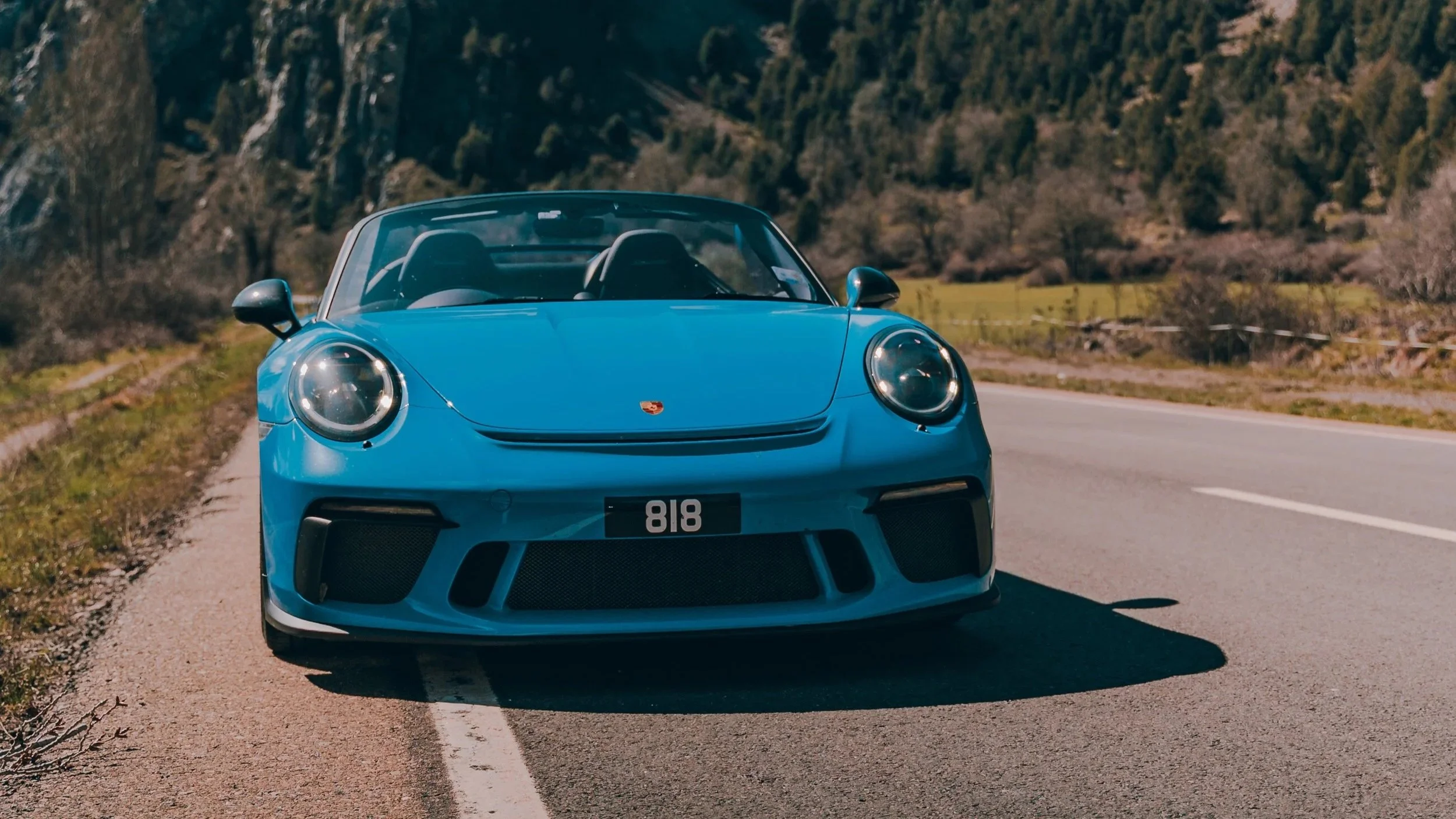 A blue Porsche 718 sports car driving on a rural road with trees and hills in the background.