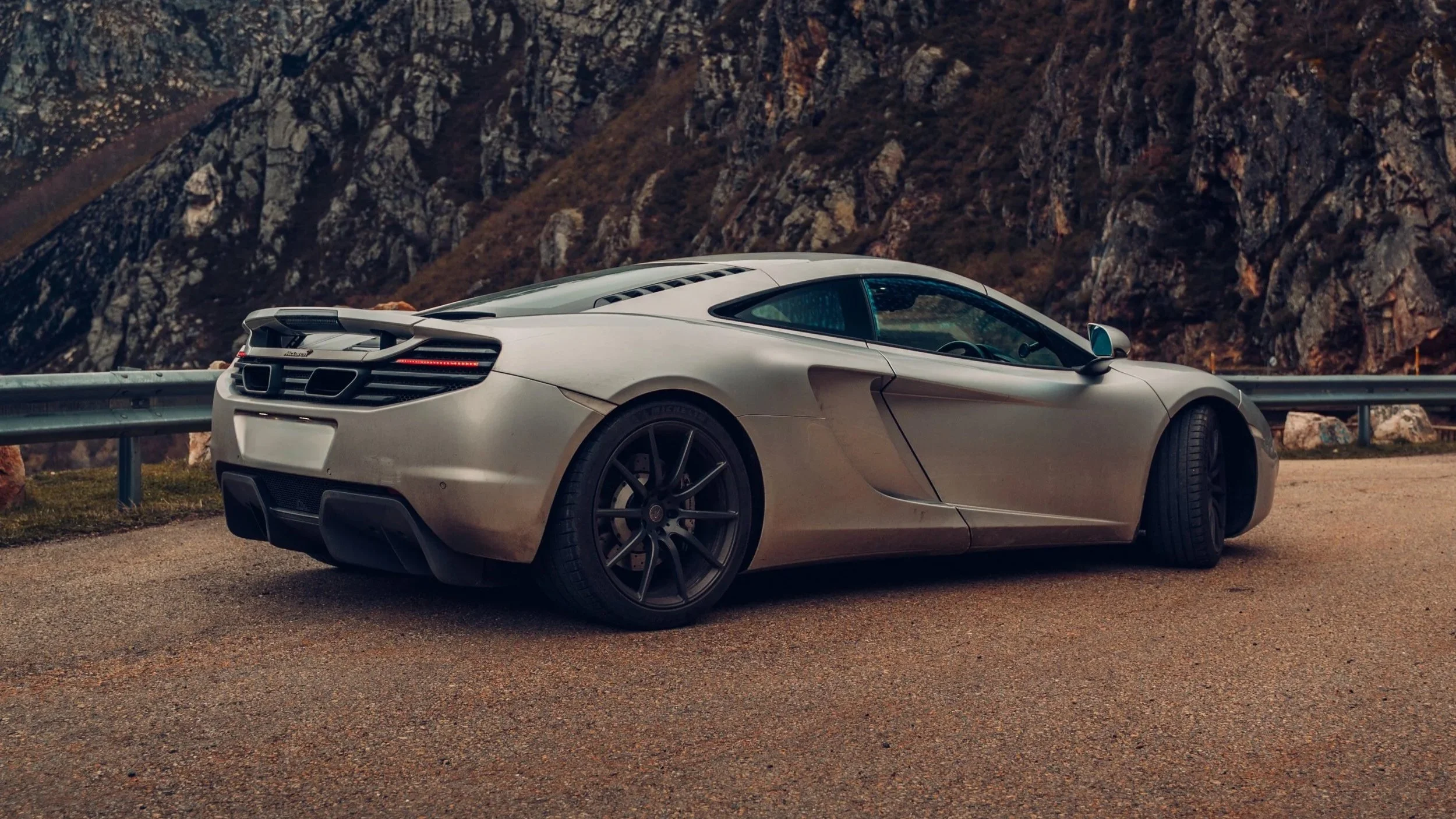 Two sports cars parked on a mountain road against a backdrop of mountains and cloudy sky. The car on the left is red with a white stripe, and the car on the right is turquoise.