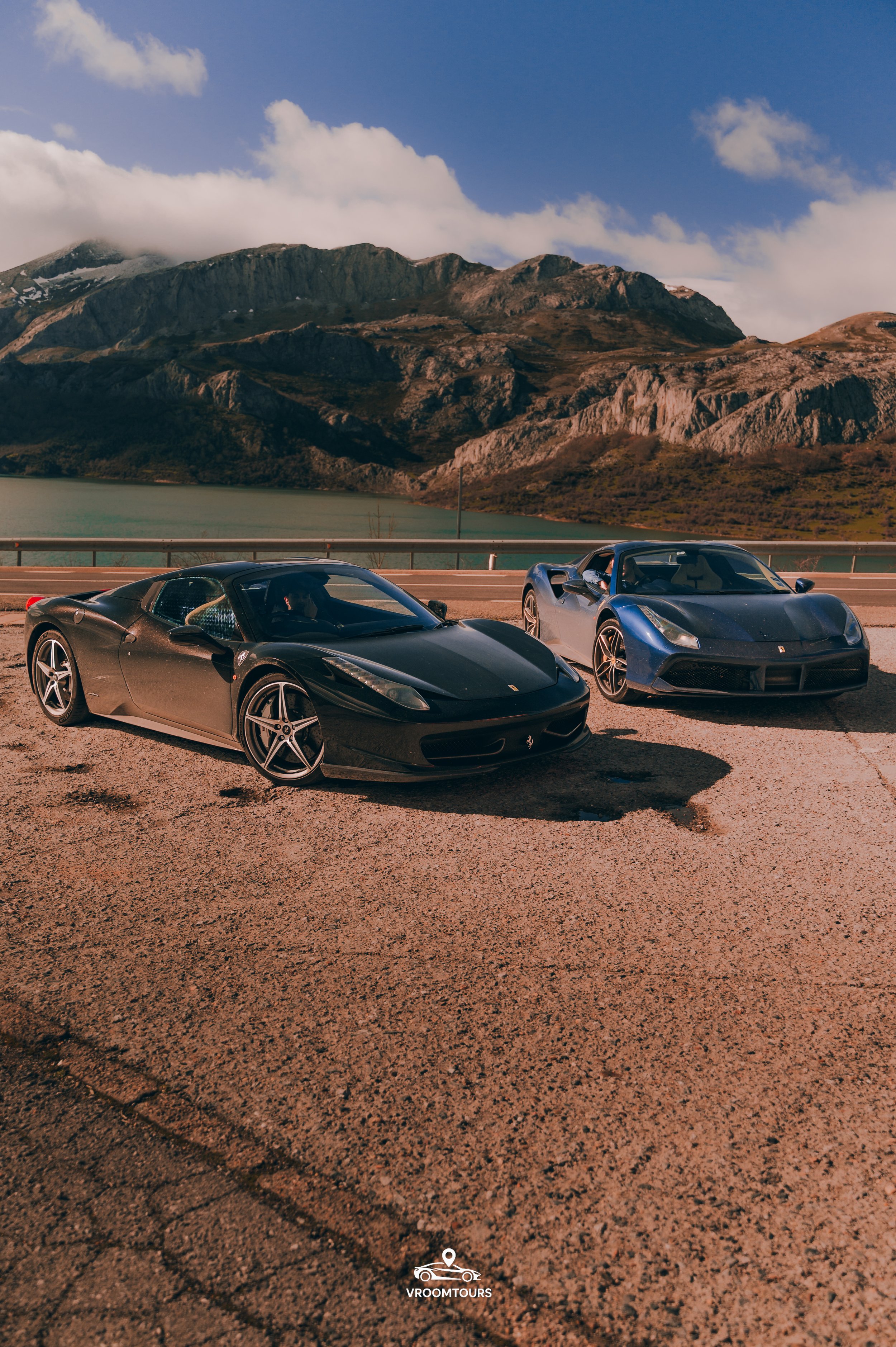 Two sports cars parked on a mountain road against a backdrop of mountains and cloudy sky. The car on the left is red with a white stripe, and the car on the right is turquoise.