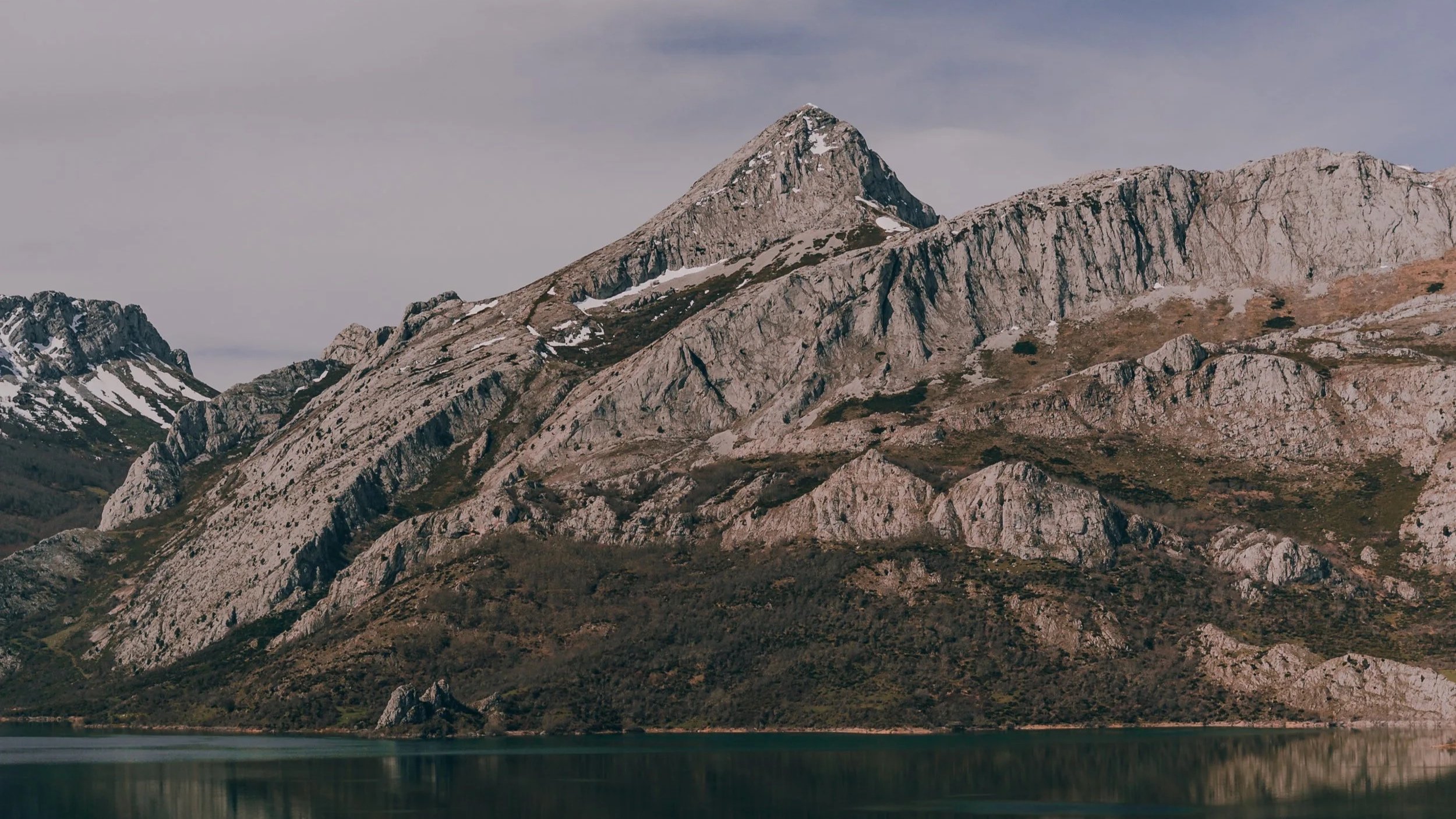 Mountain range with rugged, rocky peaks and patches of snow, next to a calm body of water.
