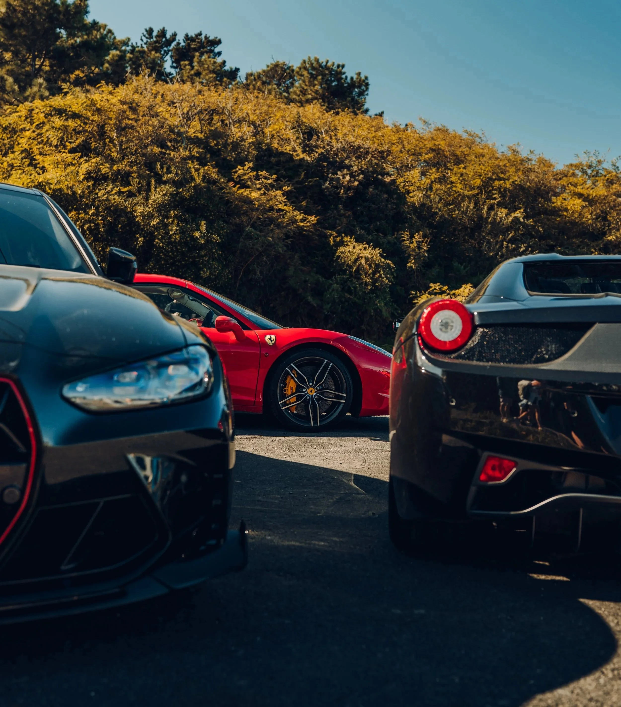 Close-up view of several luxury sports cars, including a red Ferrari and a black car, parked outdoors near a hillside with trees during daytime.