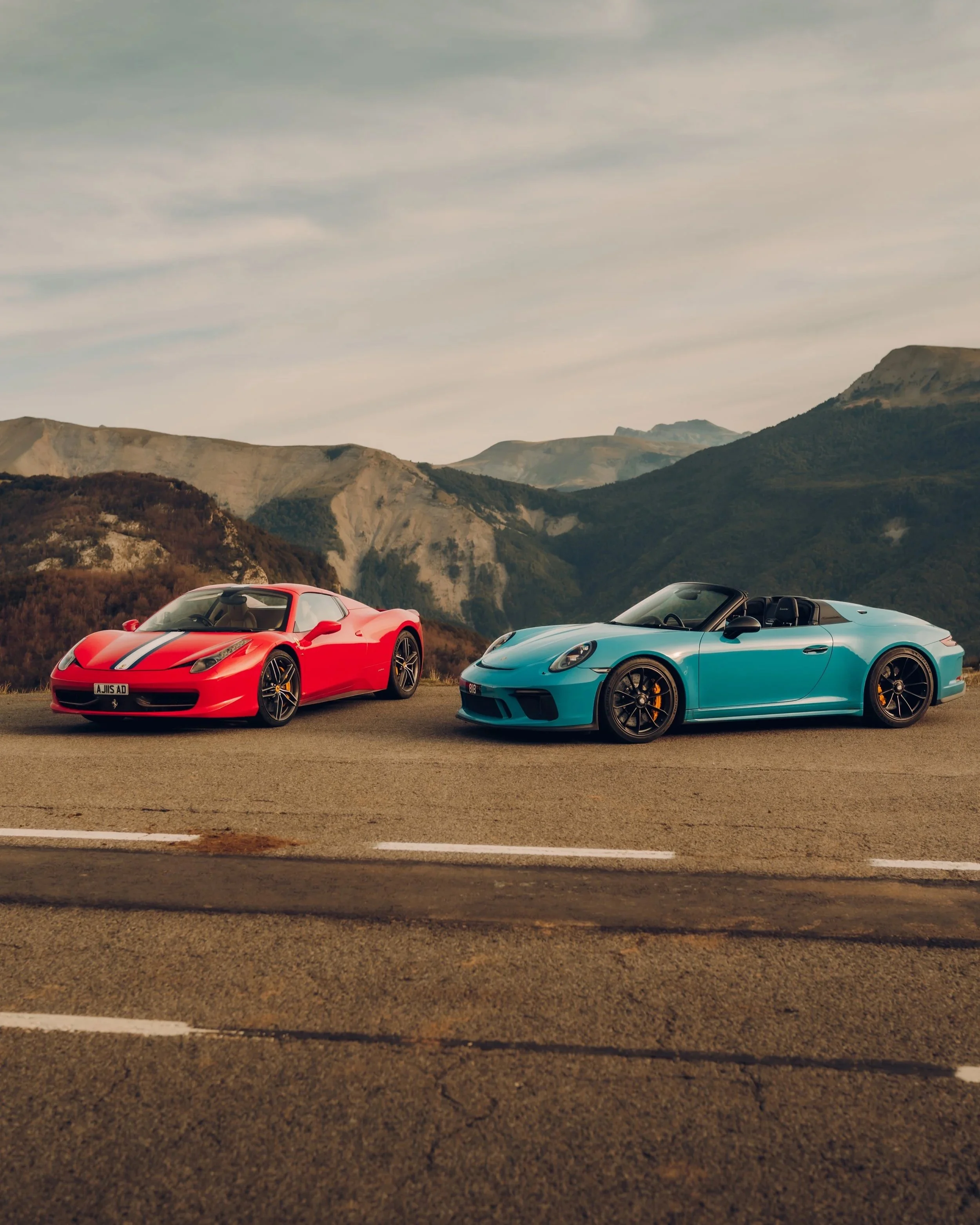 Two sports cars, a red and a blue convertible, parked on a mountain road with mountains and cloudy sky in the background.