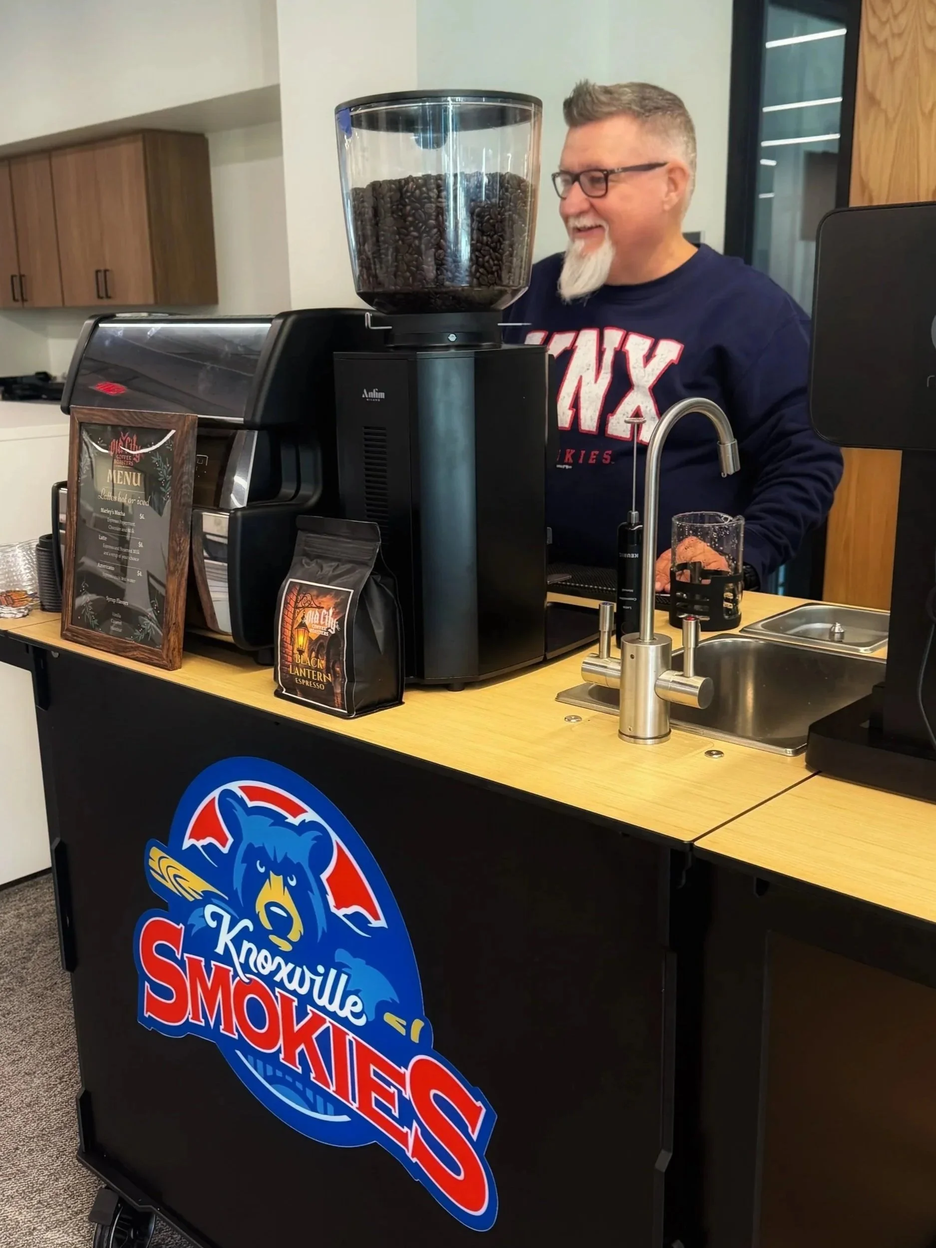 A man with glasses and a goatee making coffee at a Knoxville Smokies themed coffee stand, with a large coffee grinder filled with coffee beans and a sign menu in the background.