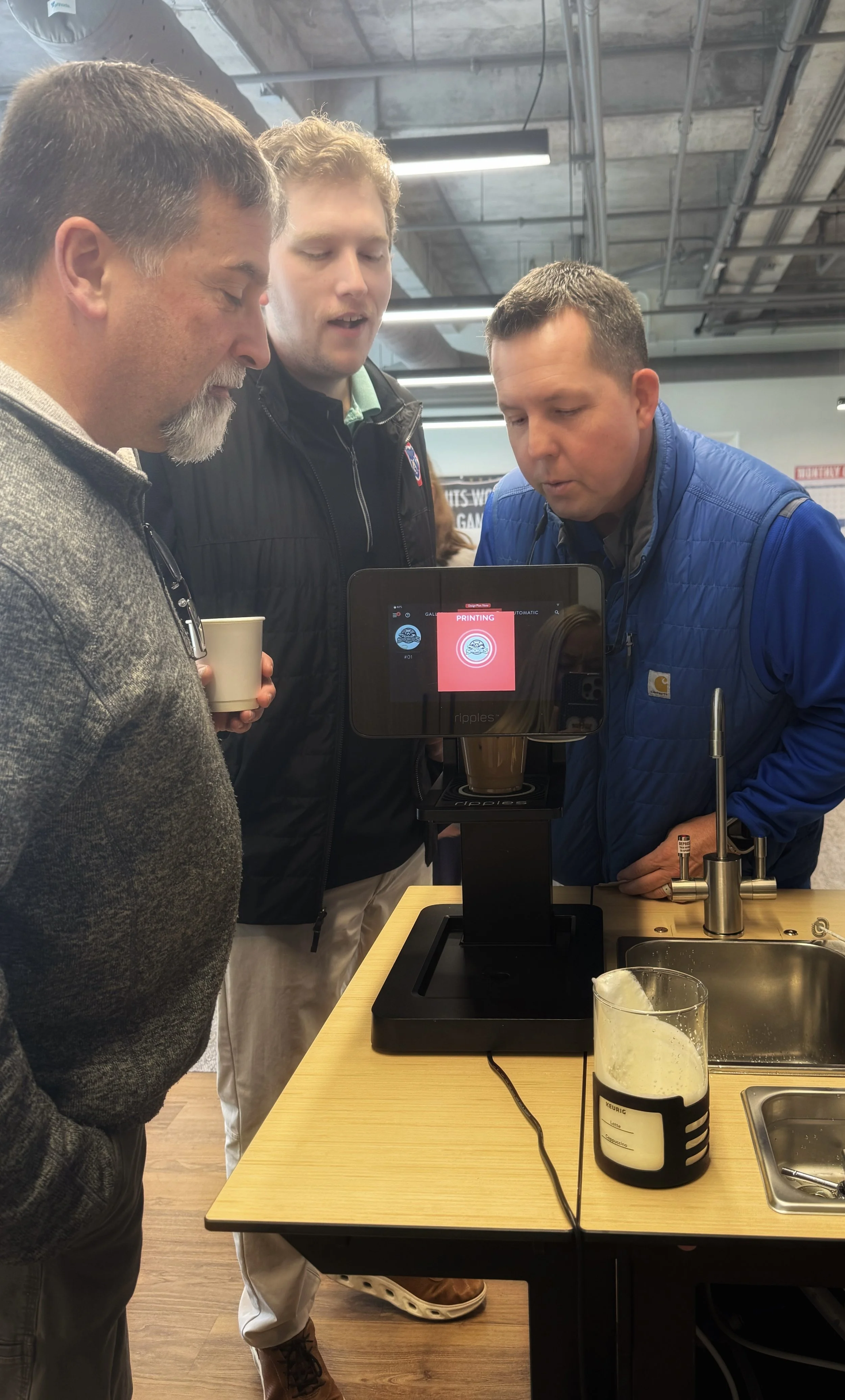 Three men are gathered around a coffee making machine with a tablet displaying the word 'PRINTING'. They are in an industrial-style room with exposed ceiling pipes and bright lighting, and they are closely observing the coffee machine.