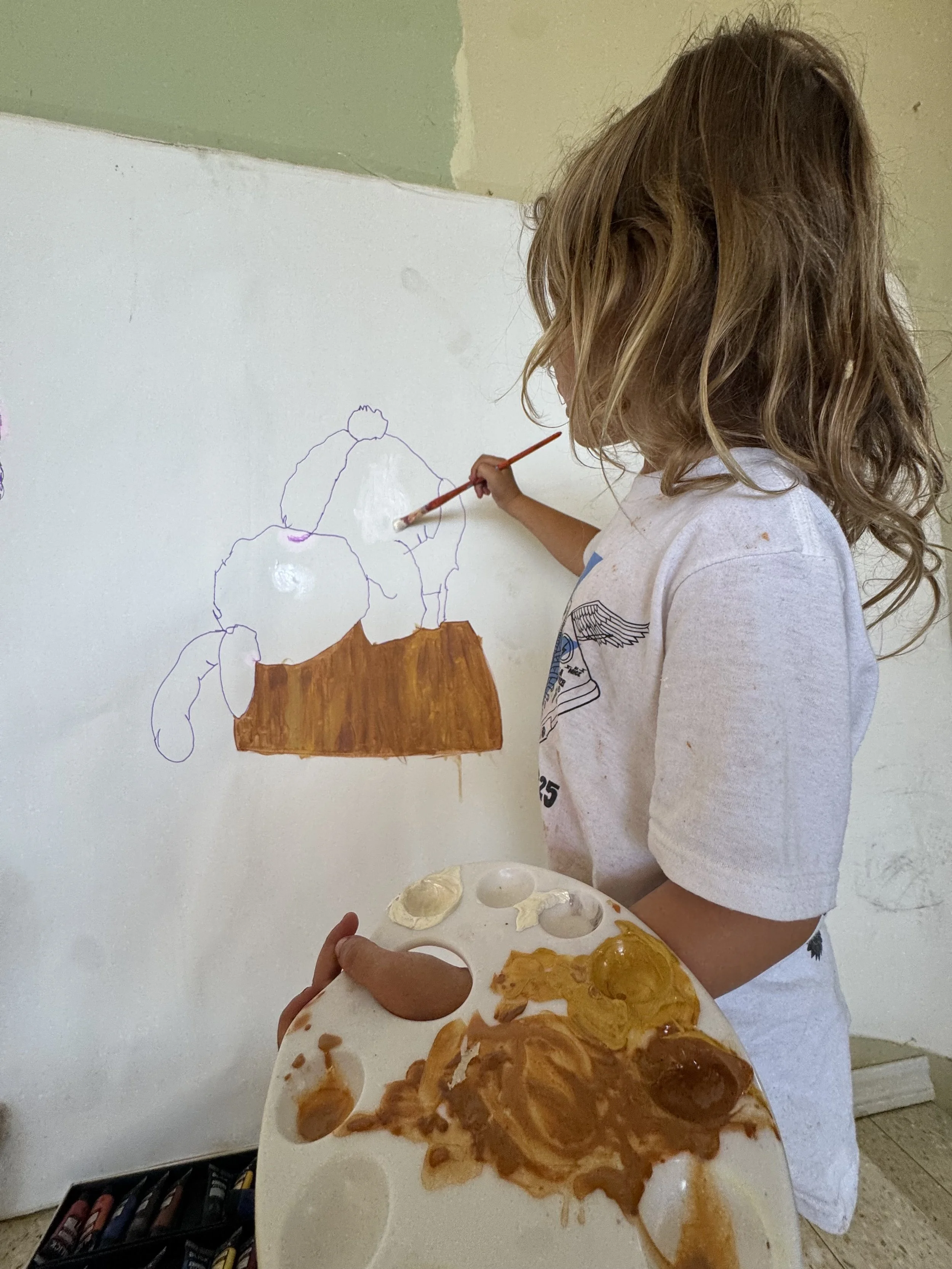 A young girl with wavy brown hair painting on a white wall with brown and purple paint. She holds a palette with mixed colors and uses a paintbrush to add details, working on an outline of a cartoon animal.