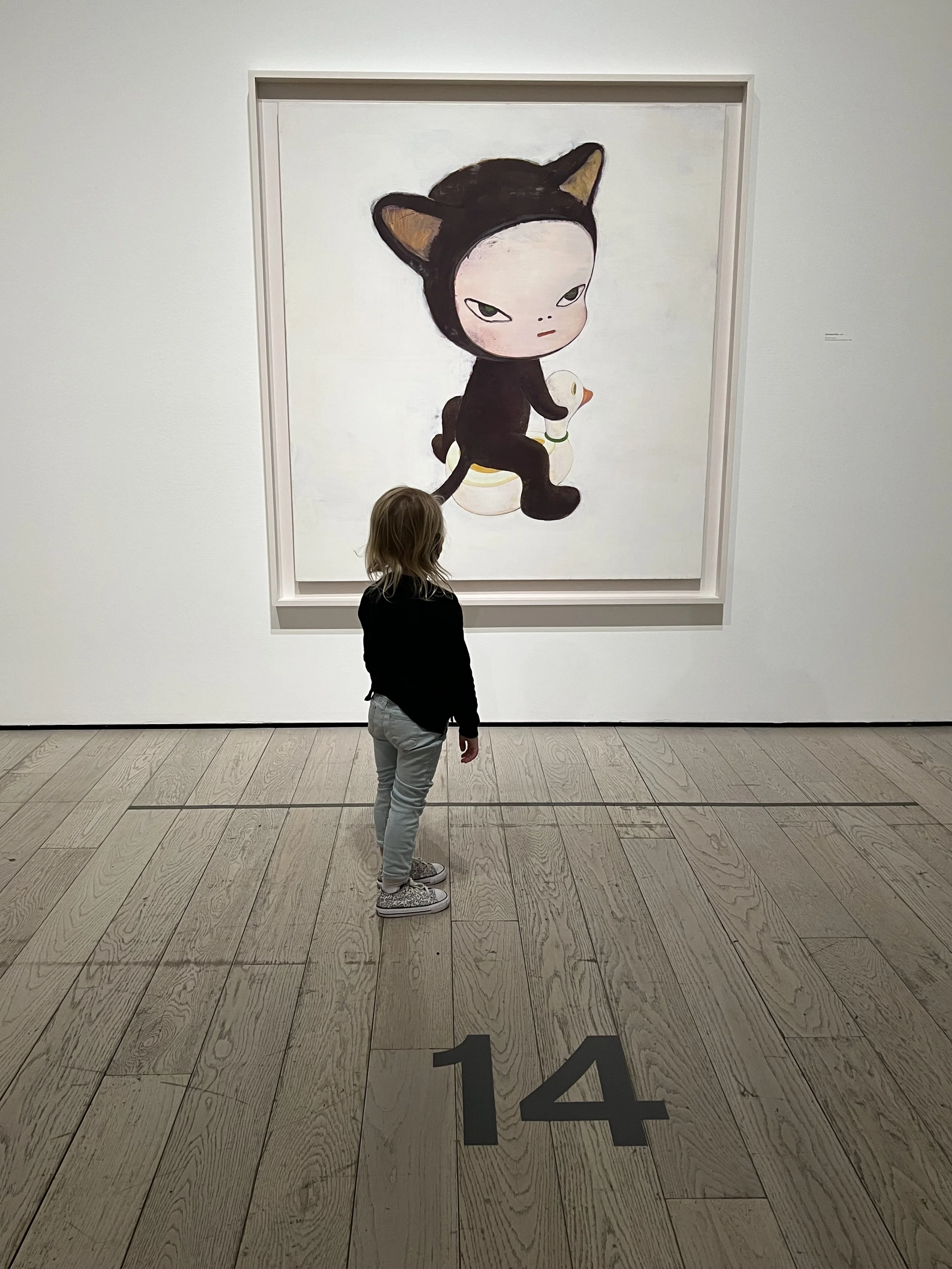 A child standing in front of a large painting of a cartoon character with a cat costume in an art gallery. The floor has the number 14 in black.