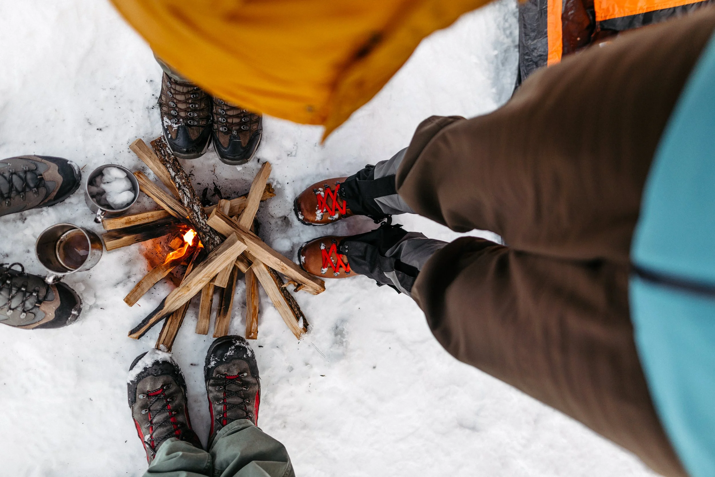 Six people standing in a circle around a small campfire on snow, wearing winter boots, with cups in hand.