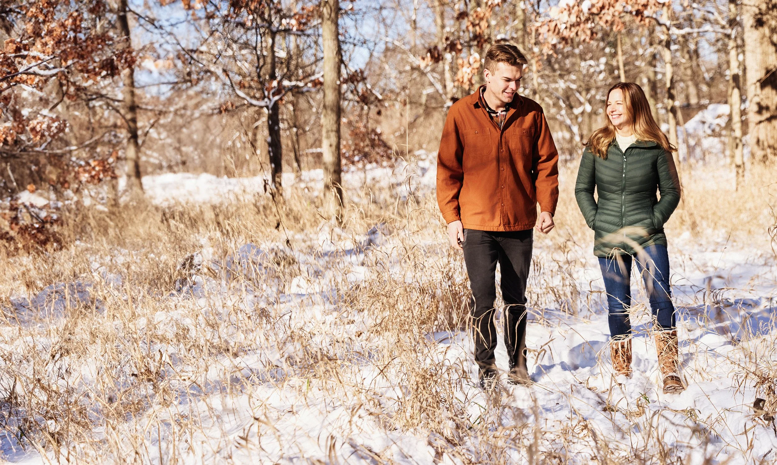 A young man and woman walking through a snow-covered field in the woods on a sunny winter day, smiling and chatting.