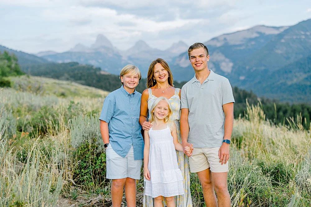 A woman with three children, standing outdoors in a grassy field with mountains in the background, smiling at the camera.