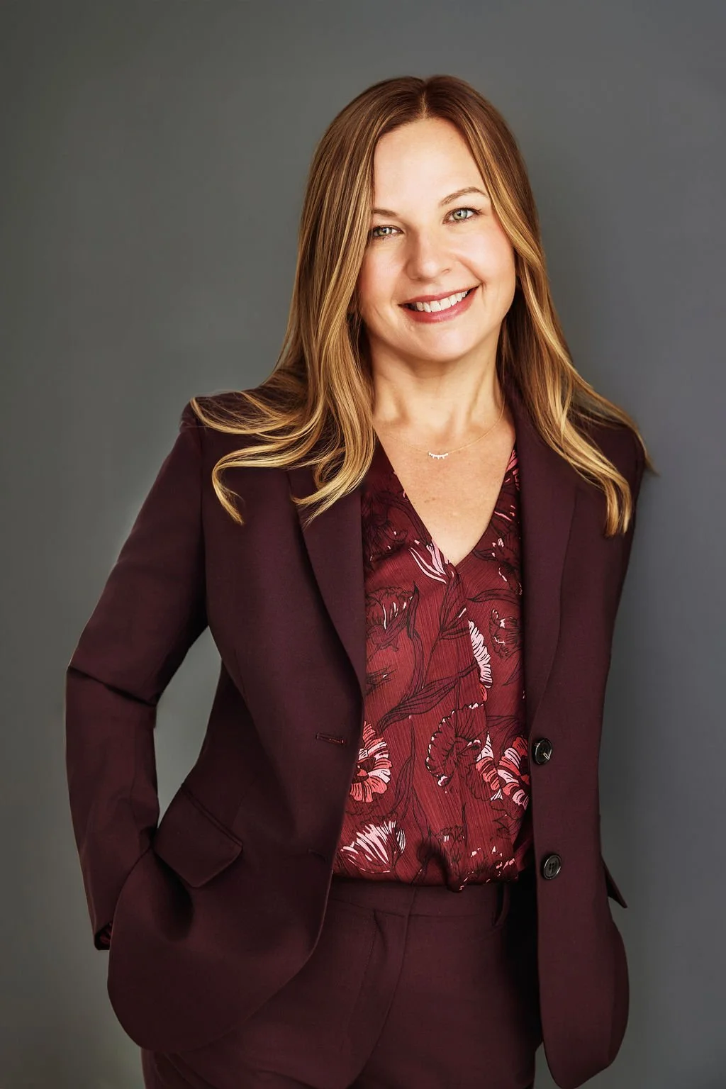 A woman with long, blonde hair wearing a deep purple blazer standing against a gray background.