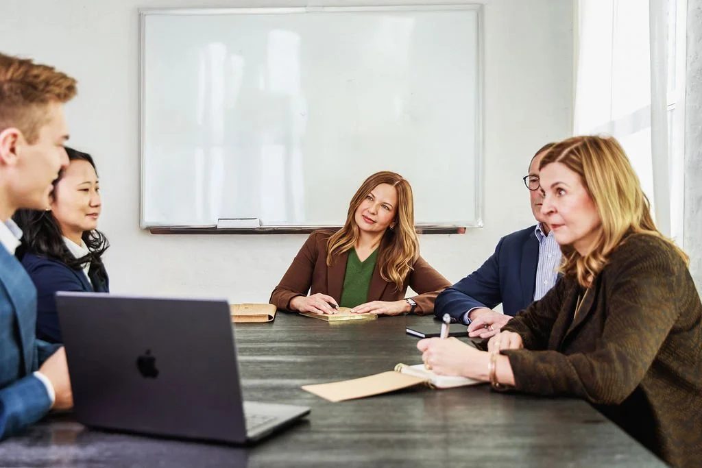 A group of five people sitting around a conference table having a meeting, with a woman in the center listening.
