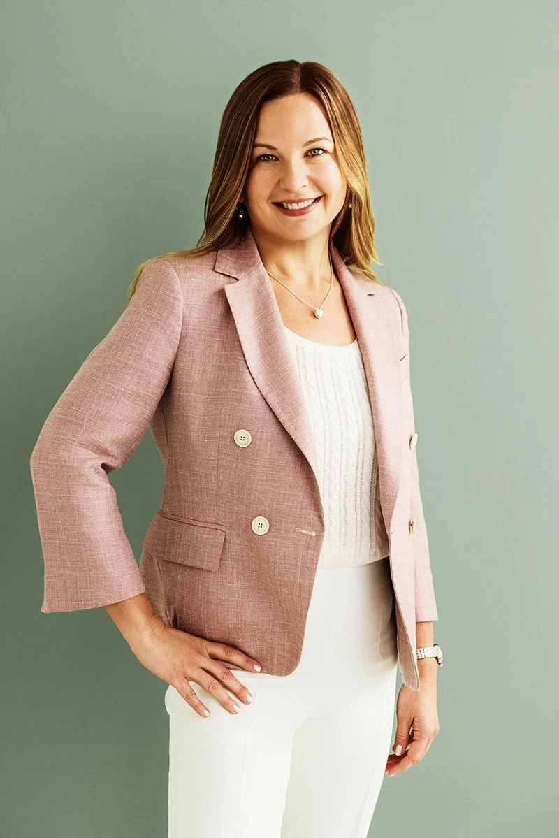 A woman with brown hair, wearing a pink blazer over a white top and white pants, standing against a light green background.