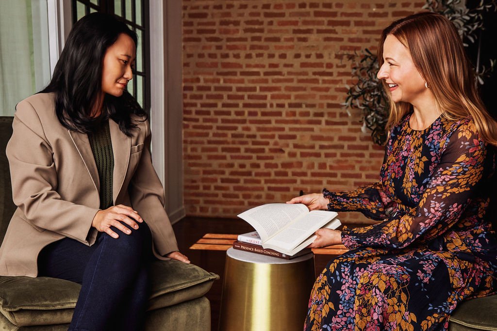 A woman with long blonde hair smiling and holding an open book sitting across from a woman in a cozy room.