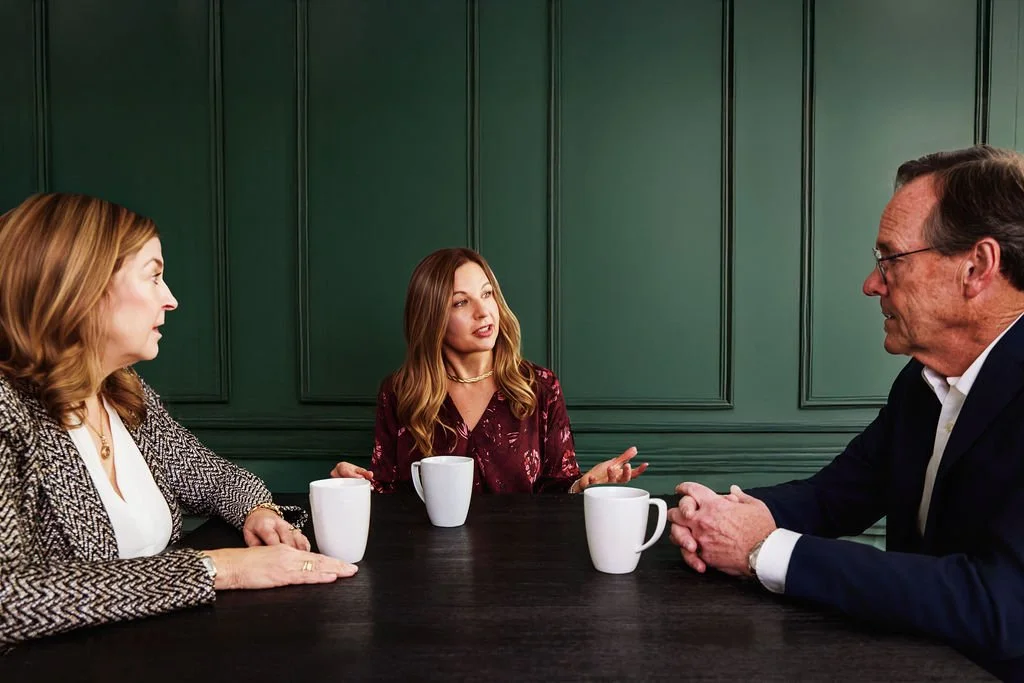Three people sitting at a dark table with white coffee mugs, engaging in conversation, against a green paneled wall.