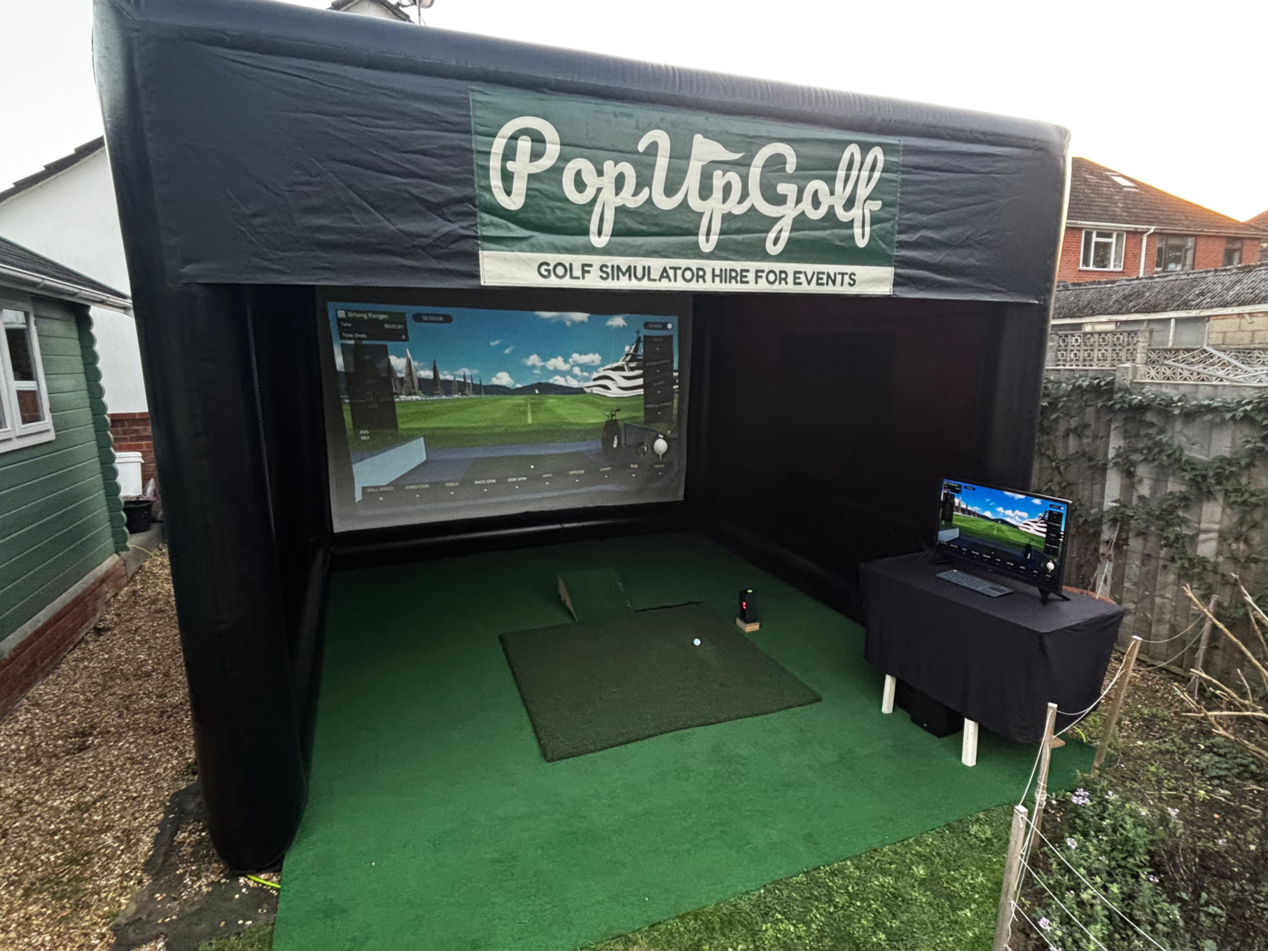 An outdoor golf simulator setup under a black tent with a sign that says 'PopUpGolf' and 'Golf Simulator Hire for Events.' It includes a large screen displaying a golf course, a smaller monitor on a table, and a virtual golf ball on a mat.