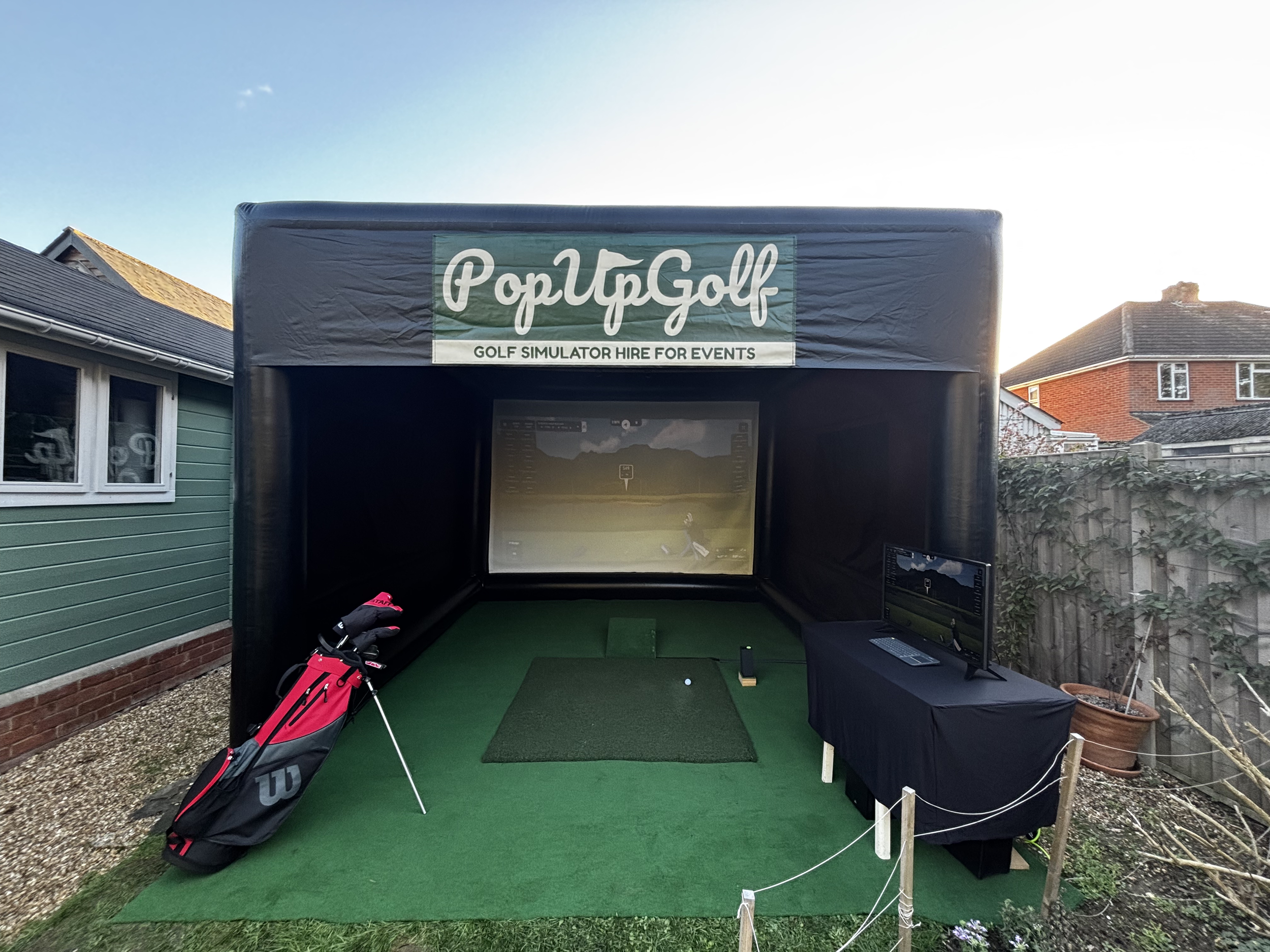 An outdoor golf simulation station labeled 'Pop Up Golf' with a large screen, golf mat, putter, and a computer setup on a table, set in a backyard with a green house and neighboring houses in the background.