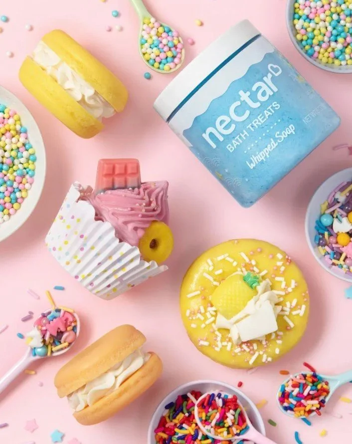 Colorful bath products and sweets on a pink background, including bath soap, sprinkles, cupcakes, and cookies.