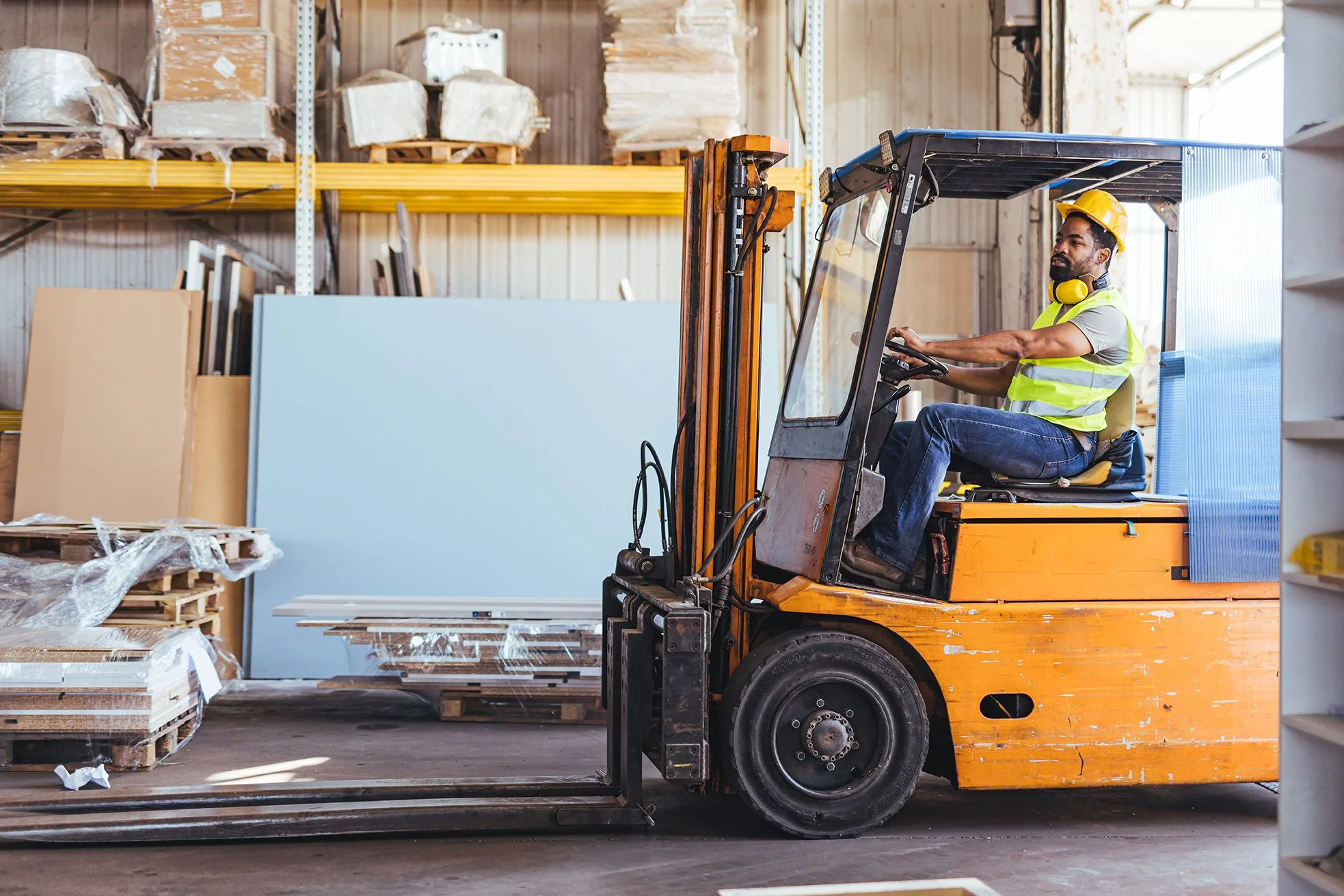 A man operating an orange forklift in a warehouse, wearing a yellow hard hat, a yellow safety vest, and hearing protection, with shelves of boxes and materials in the background.