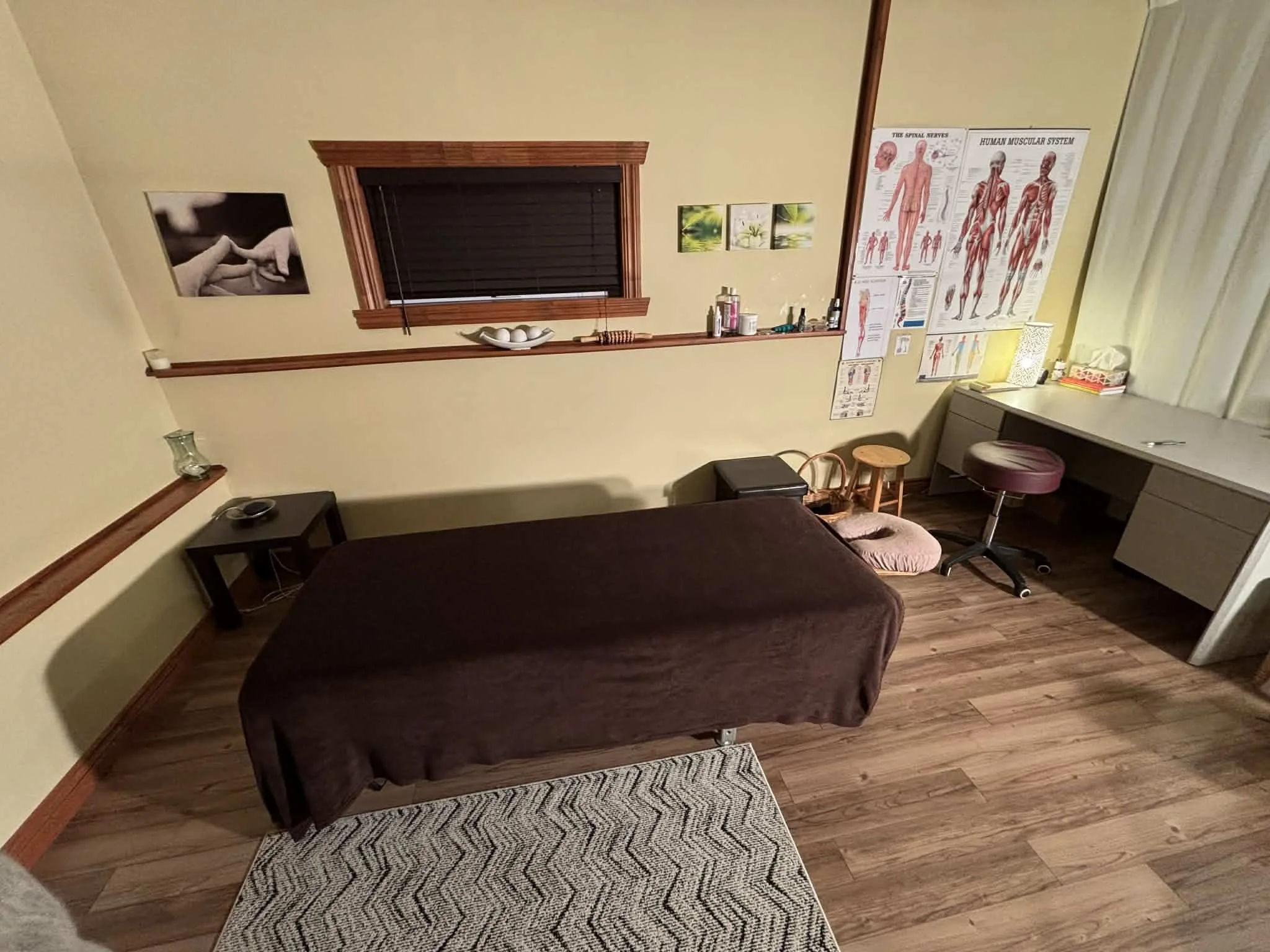 Salle de massage ou de thérapie avec un lit, un bureau avec des objets, posters anatomiques, et un petit espace de repos avec un tapis et des coussins.