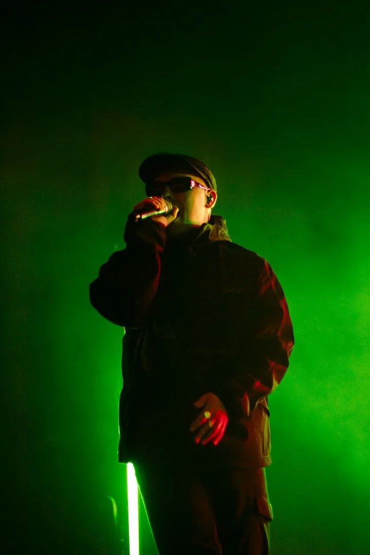 Performer on stage wearing sunglasses, holding a microphone, with green stage lighting.
