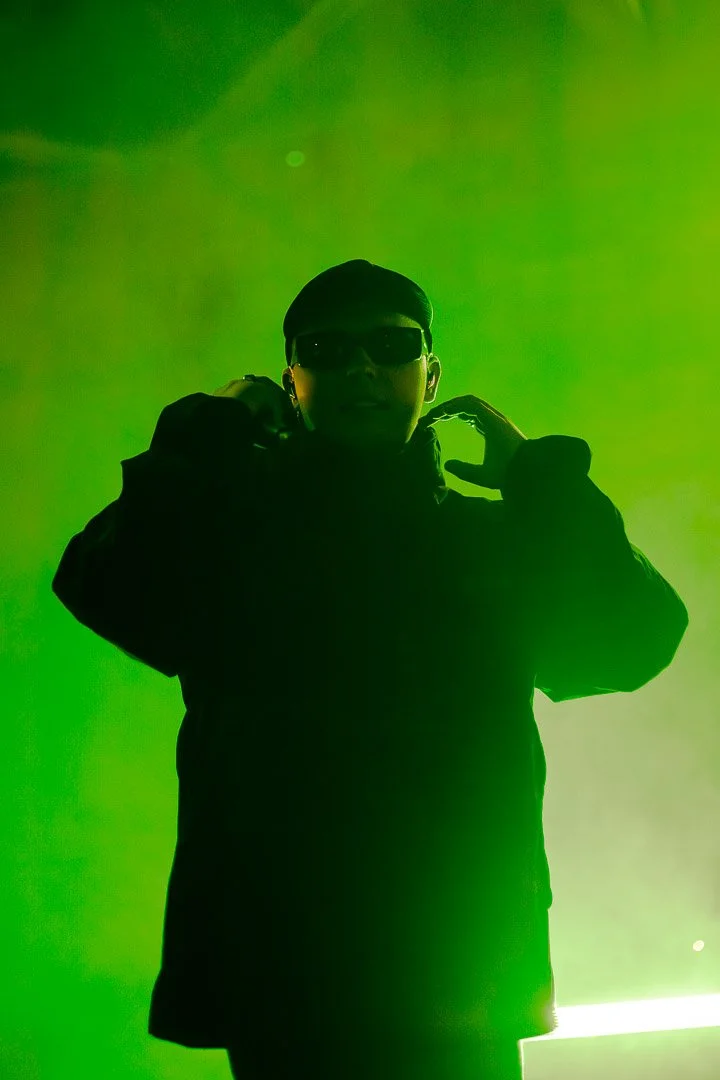 Person wearing sunglasses and a dark jacket, making a hand gesture against a green background.