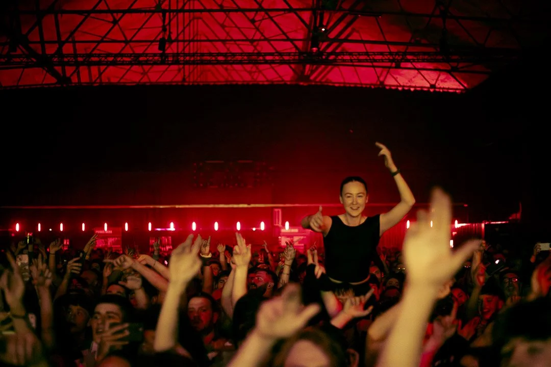 Crowd of people at a concert dancing and having fun, with a woman on someone's shoulders smiling and making a dance gesture, under red stage lighting.
